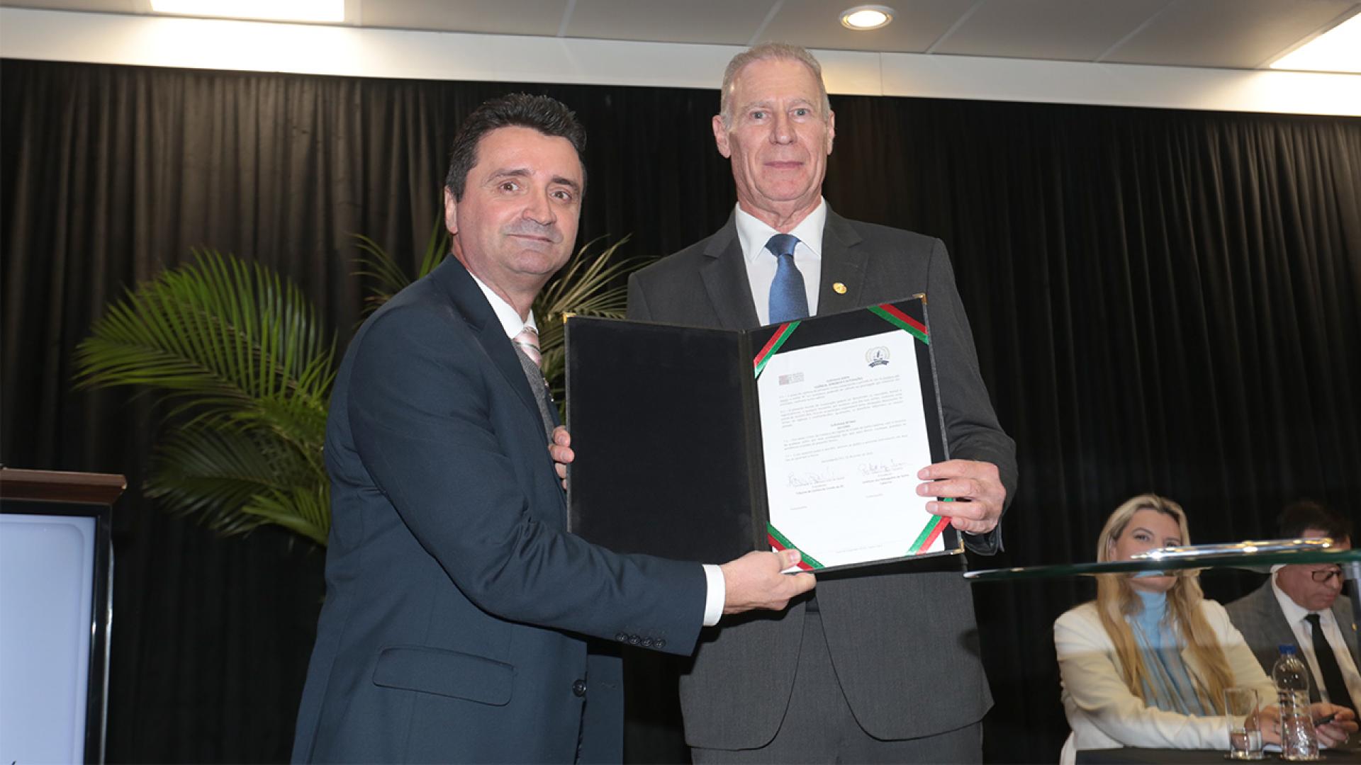 Foto do presidente do TCE/SC, conselheiro Herneus De Nadal, ao lado do presidente do IASC, Gilberto Lopes Teixeira, durante sessão solene. Eles seguram, juntos, e mostram para a foto um documento assinado. Ambos vestem terno e gravata.