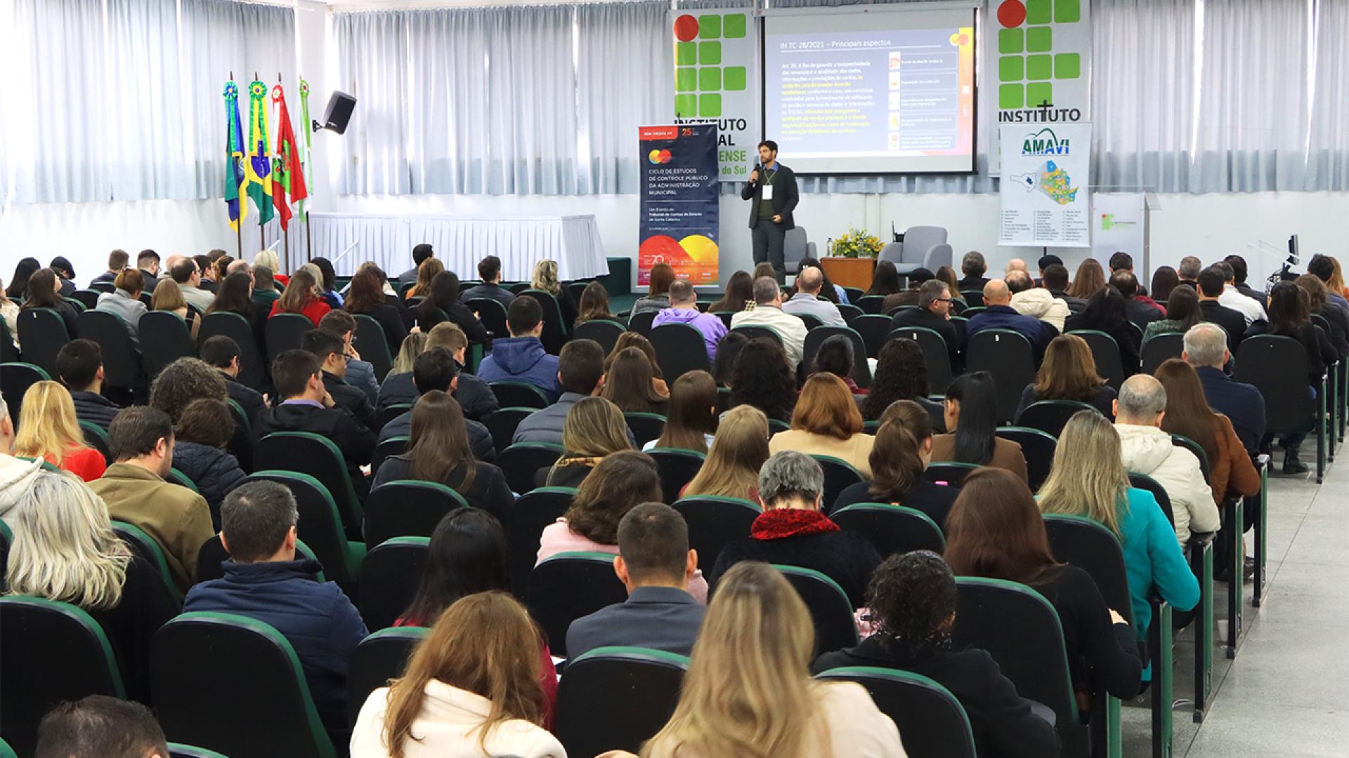 Foto mostra um homem falando ao microfone em palco que aparece ao fundo da imagem. Em primeiro plano aparecem, de costas, participantes do evento