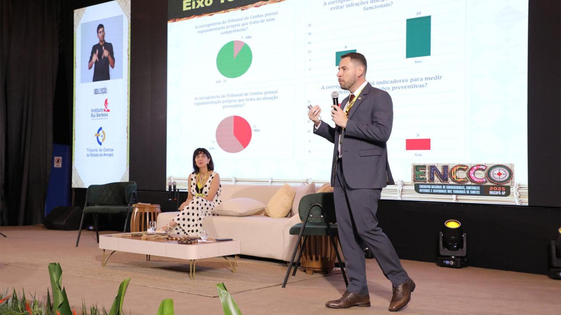 Foto horizontal do auditor fiscal de Controle Externo Geovane Cardoso durante apresentação no ENCCO 2025. Ele é um homem branco, de cabelos escuros. Veste terno e gravata e está em pé, no palco, ao lado de uma mulher. Ao fundo, no telão, é exibida a sua apresentação. 