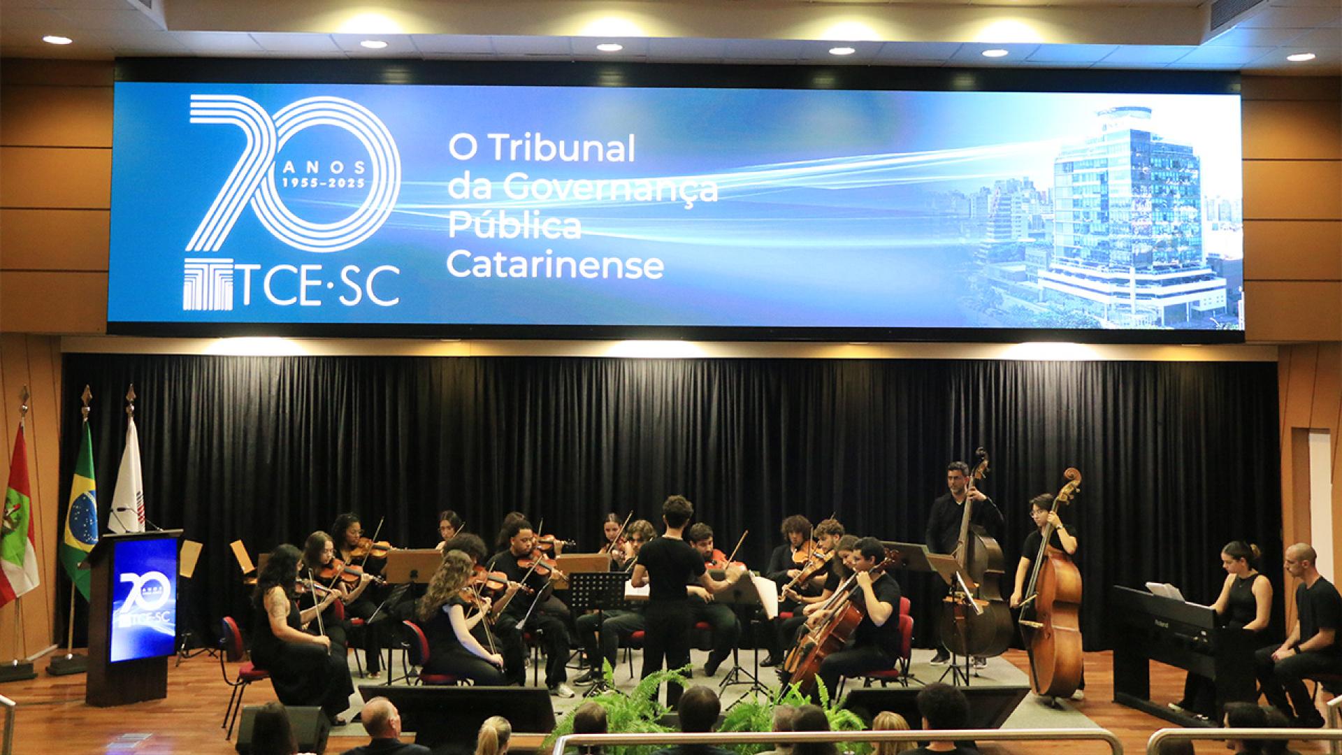 Banner horizontal com foto da Orquestra Acadêmica da Udesc no palco do auditório do TCE/SC. Há vários músicos com instrumentos. Acima deles, no telão, é projetado o logo comemorativo aos 70 anos do TCE/SC.