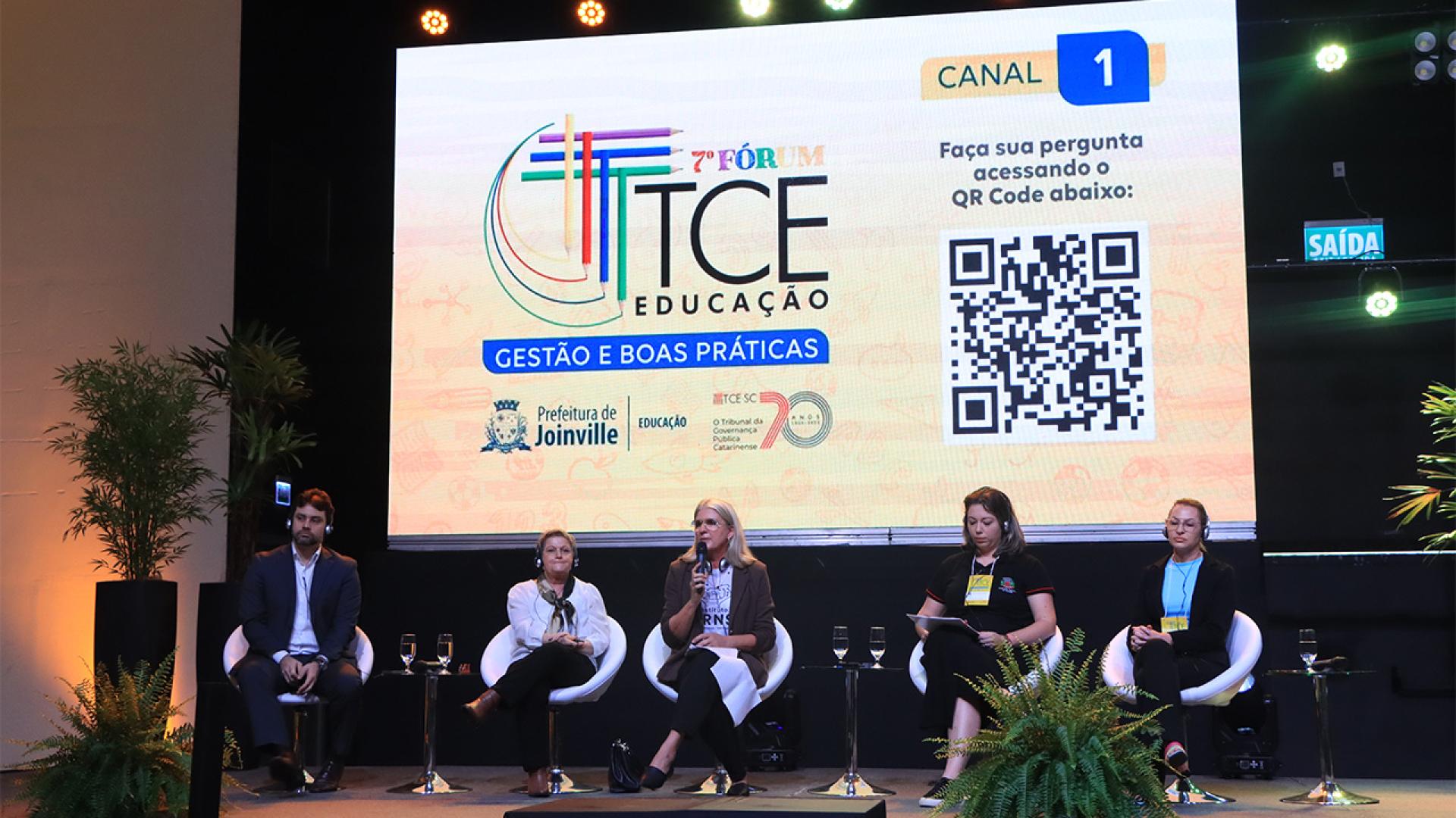 Imagem mostra cinco pessoas sentadas em poltronas brancas à frente de um telão que mostra o logotipo do Fórum TCE Educação, com predominância do tom amarelo. Há um homem, à esquerda, e quatro mulheres na imagem