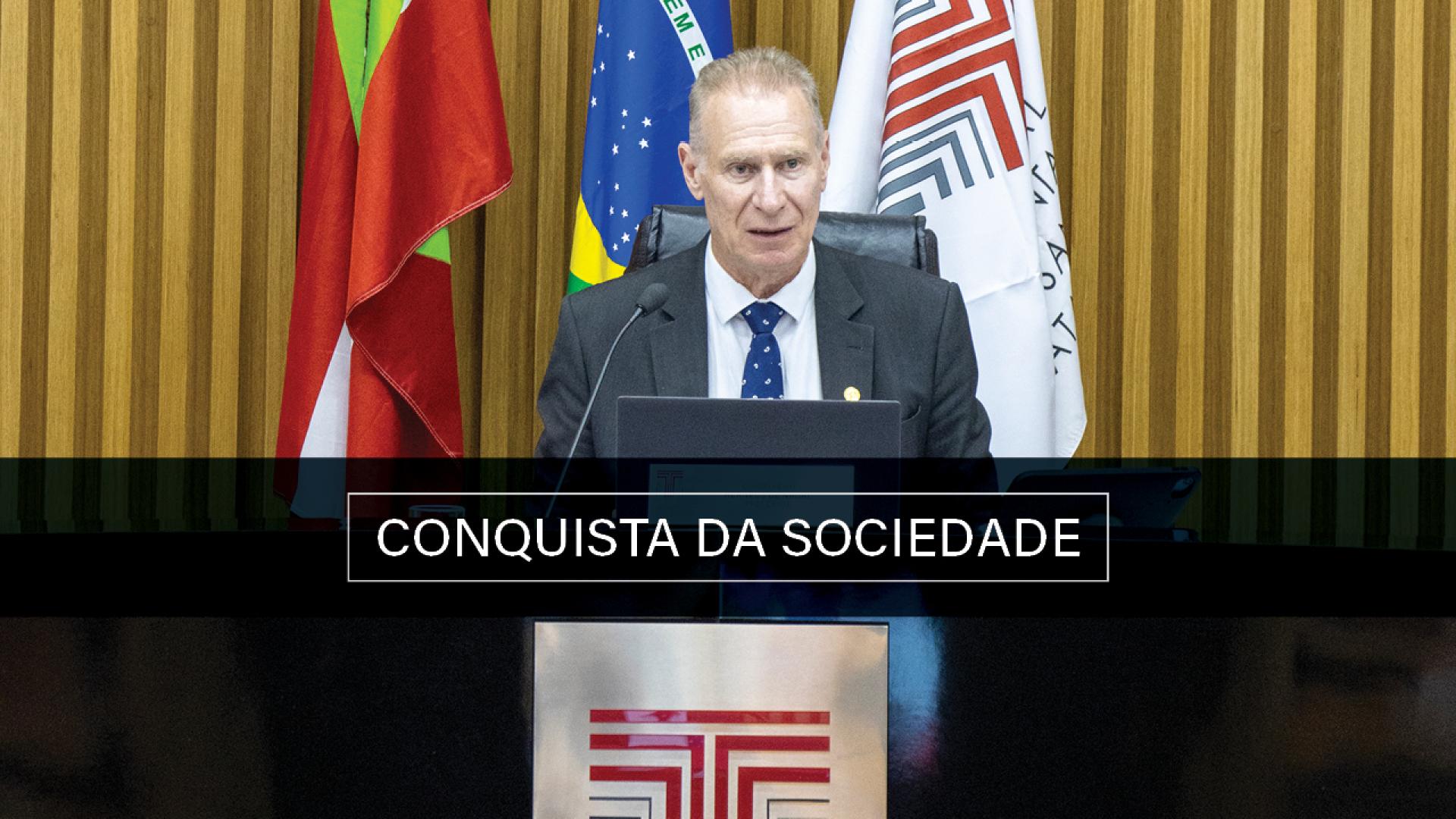 Foto colorida do presidente do TCE/SC, conselheiro Herneus De Nadal no Plenário, atrá dele as bandeiras de Santa Catarina, do Brasil e do TCE/SC ( da esquerda para a direita). Ao centro está escrito "Conquista da Sociedade". 