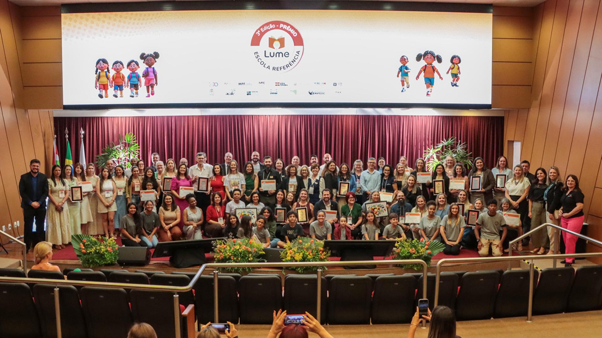 Banner horizontal com foto de várias pessoas no palco do auditório do TCE/SC. São os representantes das escolas premiadas pelo Prêmio Lume e autoridades, entre integrantes do TCE/SC, da educação do Estado e das demais instituições do acordo Lume. Alguns seguram certificados recebidos.