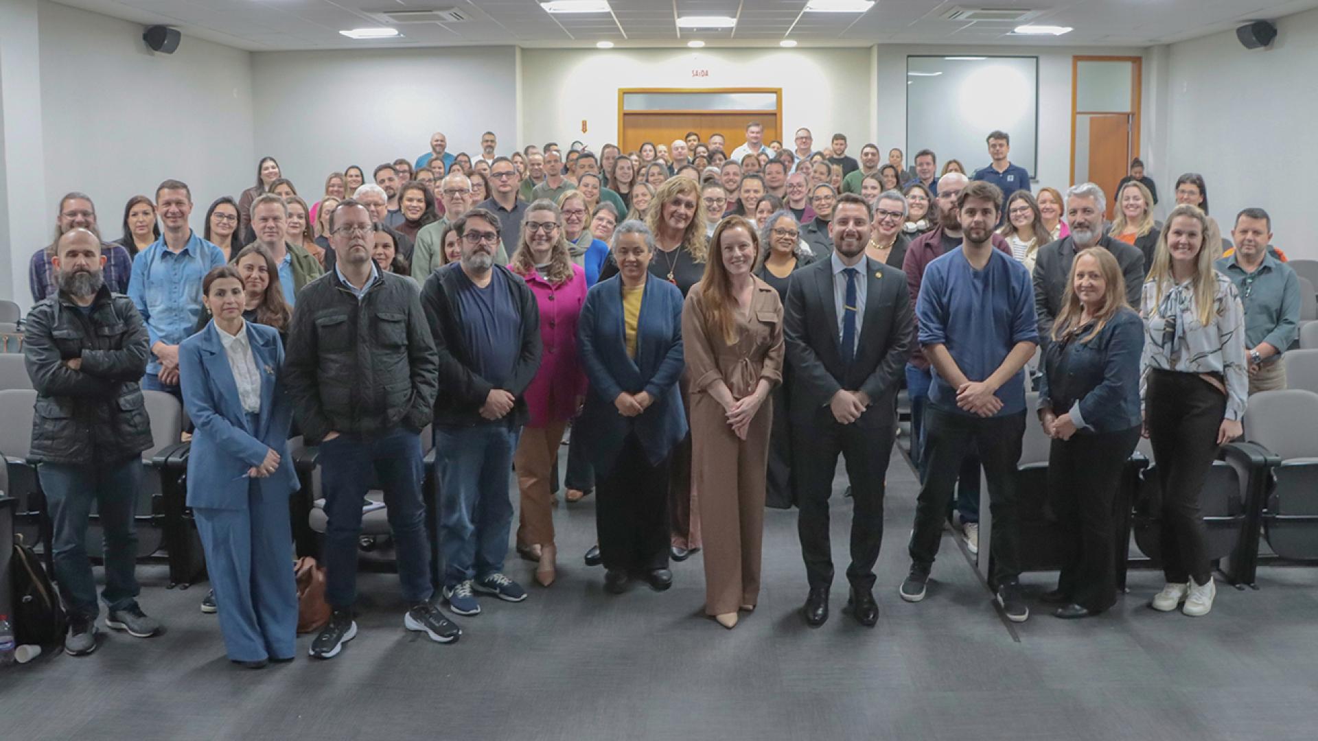 Banner horizontal com foto de um grupo em um auditório durante o Ciclo de Estudos. Dezenas de participantes estão posicionados entre as fileiras de cadeiras, voltados para a câmera. Na primeira fila, destacam-se os auditores fiscais de controle externo Vivian Savedro e Geovane Eziel, acompanhados de outros participantes do evento.