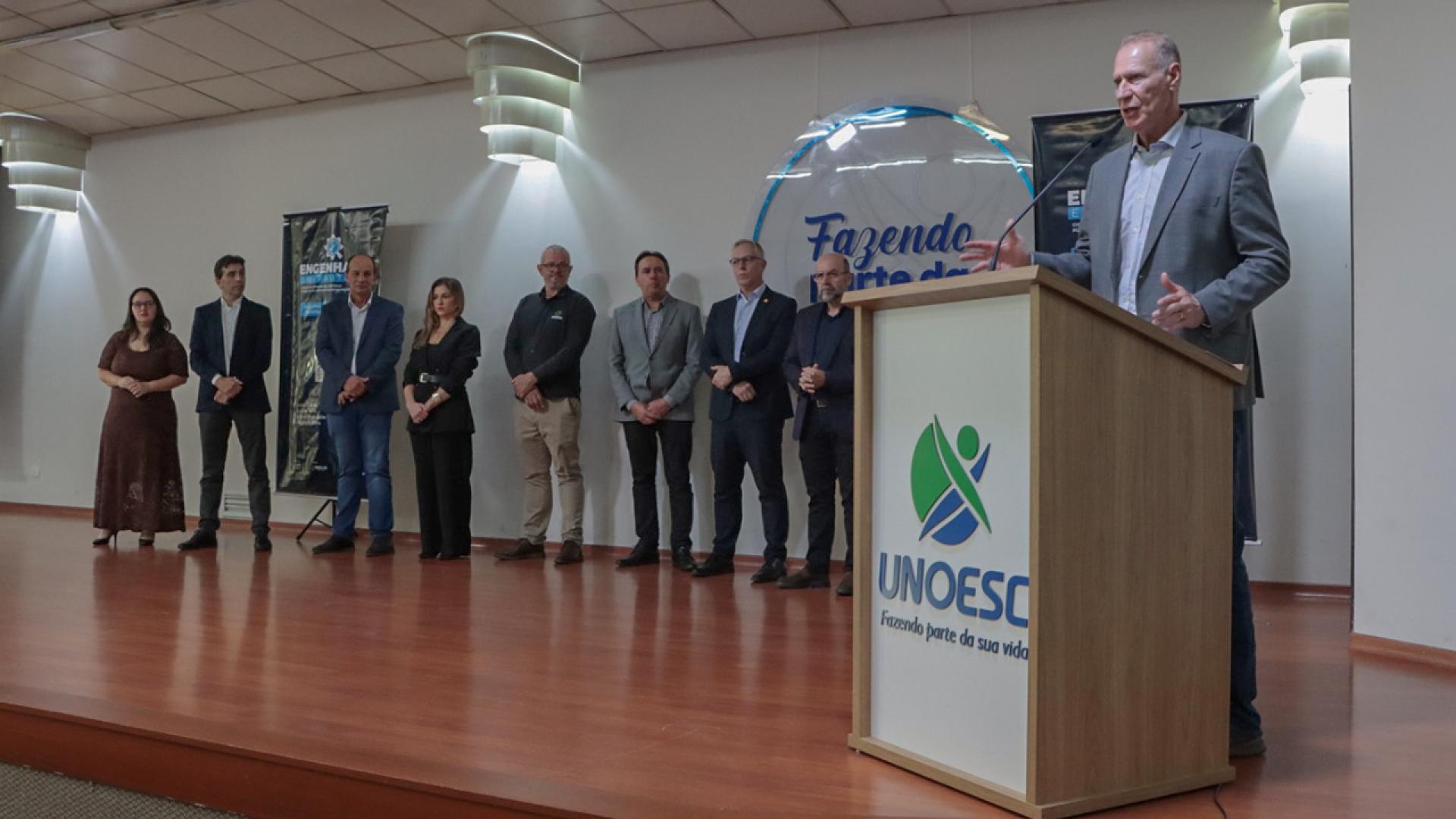 Banner horizontal com foto do presidente do TCE/SC, conselheiro Herneus De Nadal, discursando no púlpito, na abertura do evento, no auditório da Unoesc. Ele está à direita da foto. À esquerda, no palco, lado a lado, equipe do TCE/SC que o acompanha, entre diretores e servidores.