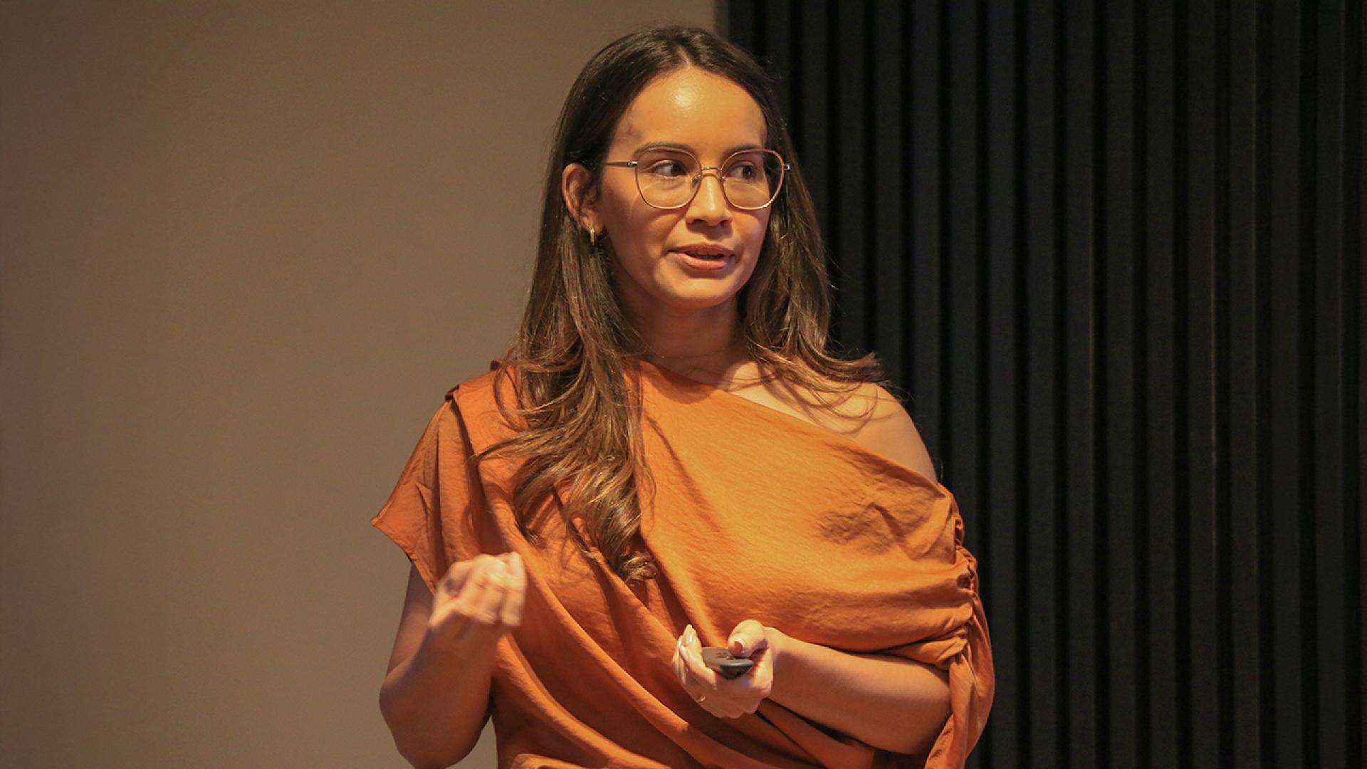 Banner horizontal com foto da servidora Julia Maria Leal dos Santos durante apresentação de mestrado. Ela utiliza óculos e veste roupa em tom terroso, segurando um controle nas mãos enquanto fala, com expressão atenta e gestual leve, em ambiente interno com iluminação focada e fundo escuro.