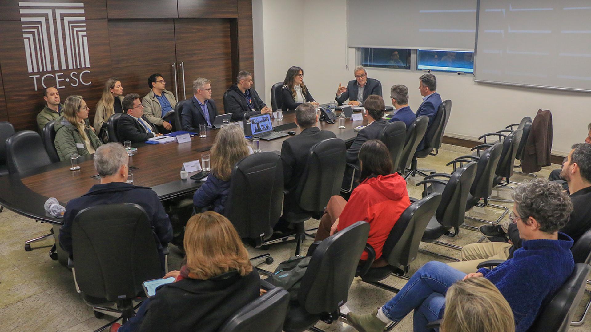 Banner horizontal com foto dos vários participantes da mesa de consensualismo reunidos no TCE/SC. Eles estão sentados diante de uma mesa, enquanto conversam. Entre os presentes, o coordenador dos trabalhos, Neimar Paludo, outros integrantes do TCE/SC, da Casan e de outras instituições envolvidas. 