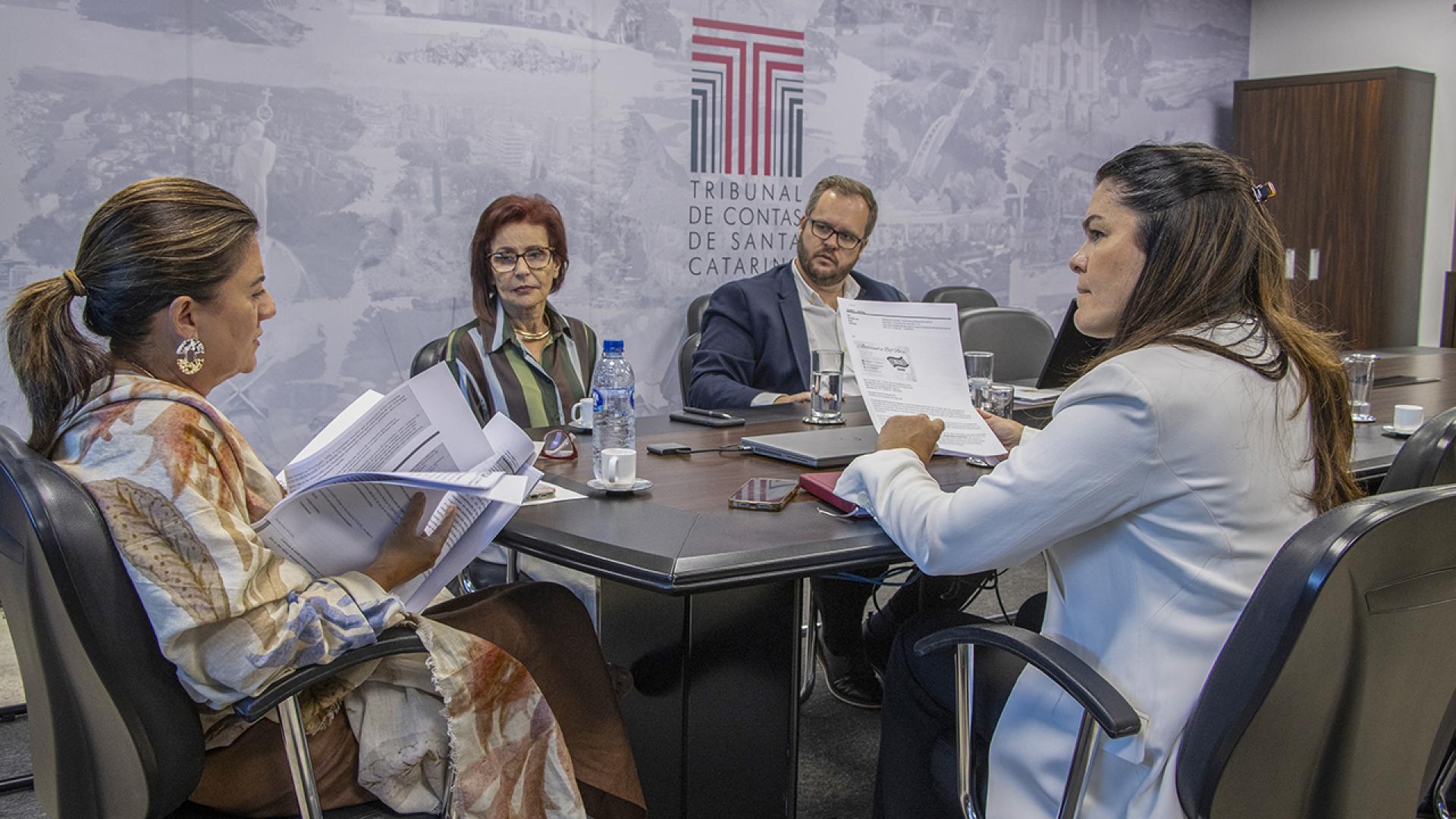 Foto horizontal de representantes da Associação dos Consórcios Interfederativos de Saúde do Estado de Santa Catarina reunidos com a chefe de Gabinete ajunta da Presidência, Raquel Zomer, e a diretora-geral adjunta de Controle Externo, Monique Portella. Eles estão sentados diante de uma mesa, enquanto conversam. Na parede, plotagem do logo do TCE/SC.
