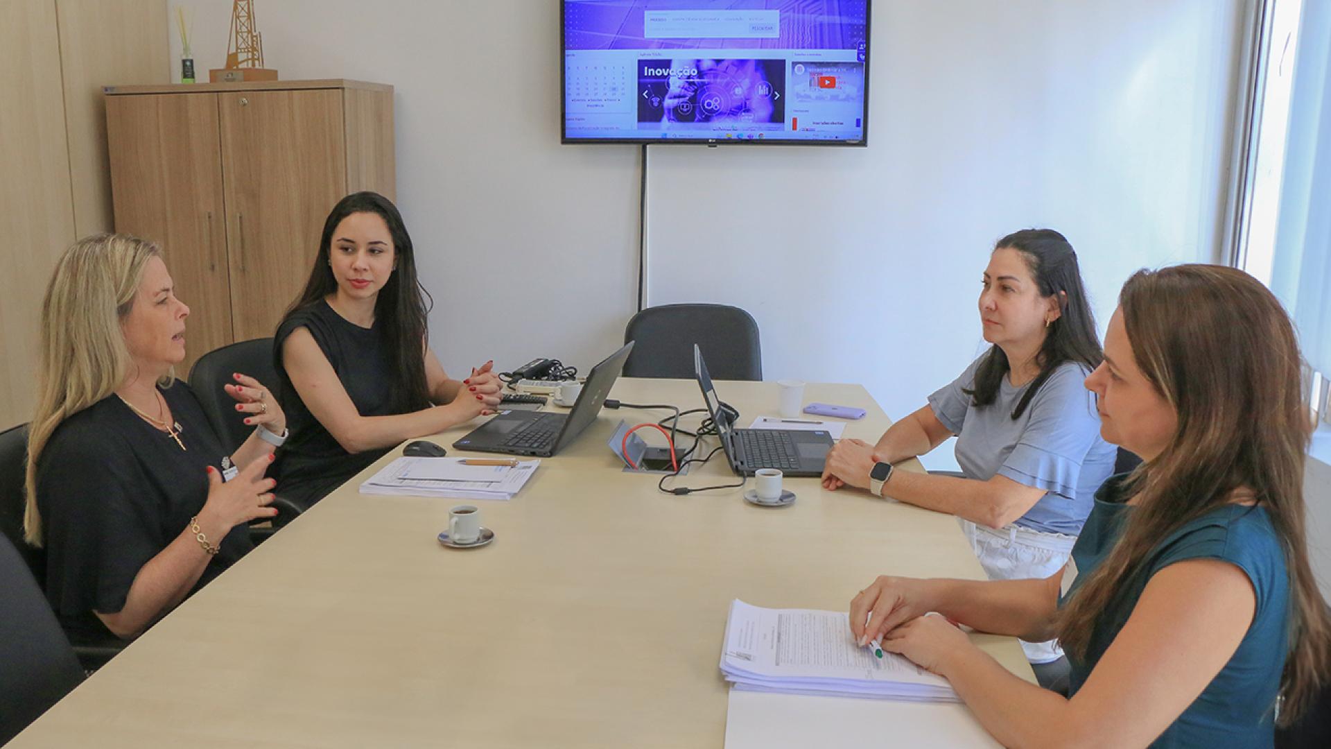 Imagem mostra quatro mulheres ao redor de uma mesa de cor clara. Ao fundo, há um monitor que mostra dados. Duas das mulheres estão com laptops abertos em sua frente