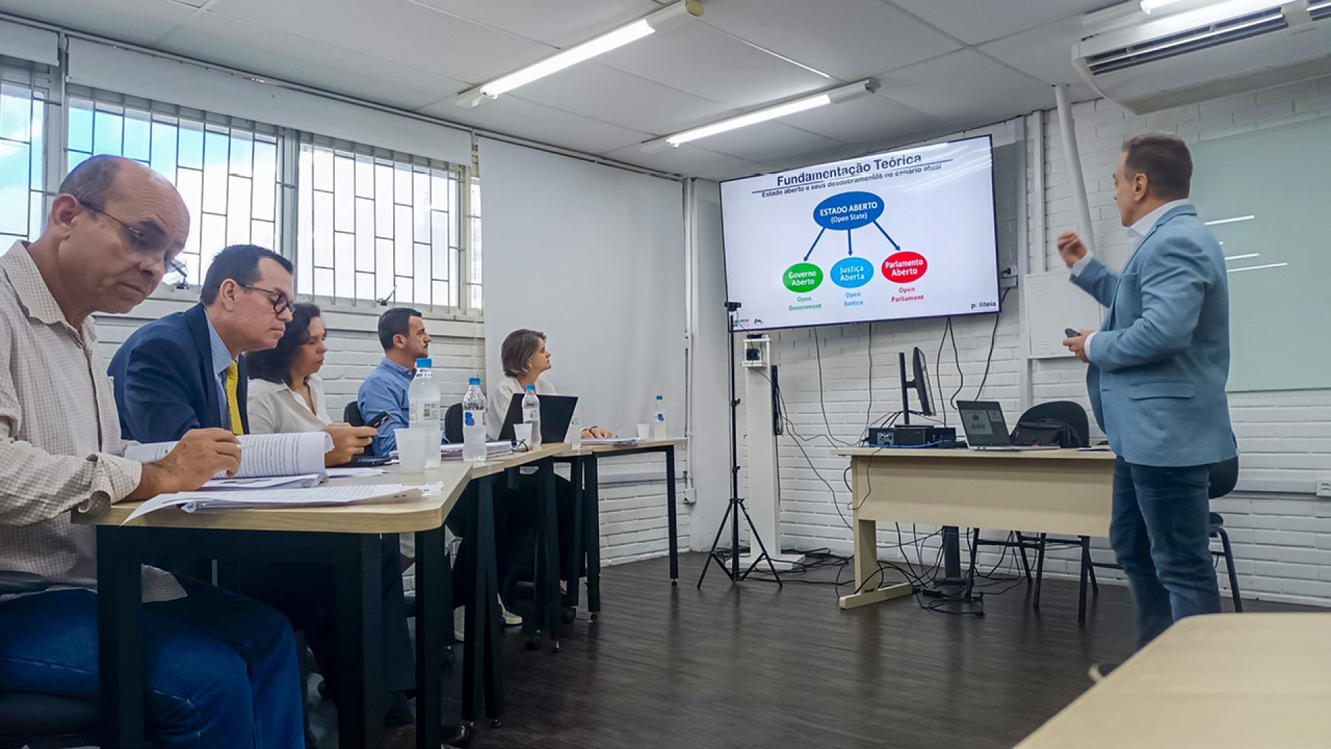 Foto horizontal do servidor Renato Costa durante defesa de tese de doutorado. Ele está à direita, em pé, apresentado. À esquerda, a banca, composta, entre outras autoridades, pelo conselheiro Adircélio de Moraes Ferreira Júnior. 