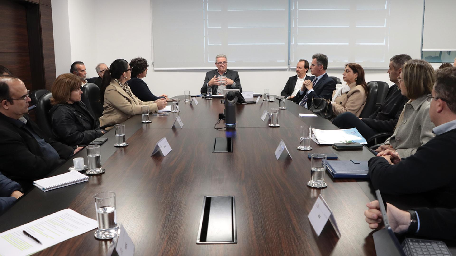 Foto horizontal que mostra os participantes da reunião sentados ao redor de uma mesa enquanto conversam. Ao centro, o coordenador da mesa de consensualismo, Neimar Paludo. Entre os demais presentes, integrantes do TCE/SC, da Casan e do MPSC, e o deputado estadual Marquito. Sobre a mesa, há papéis, canetas e copos de água. 