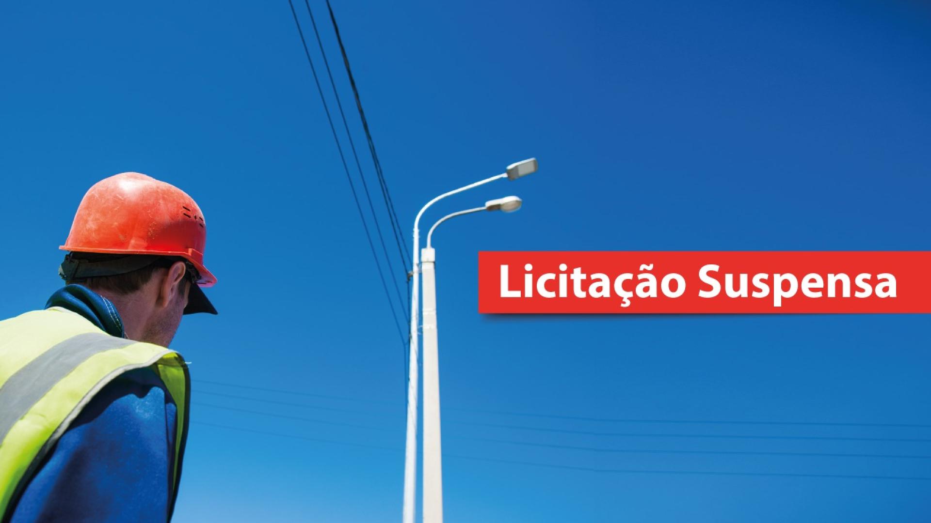 Imagem apresenta um poste com luminárias e fios elétricos sob céu azul, com texto em destaque: “Licitação Suspensa” 
