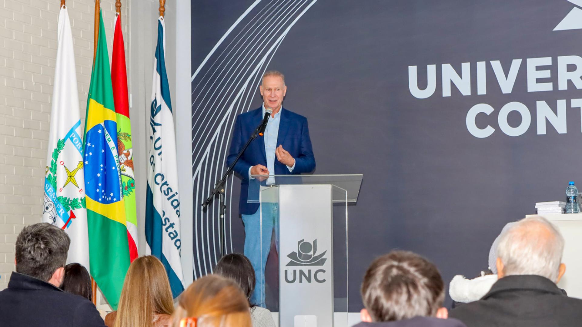 Banner horizontal com foto do presidente Herneus De Nadal discursando no púlpito, no palco do auditório da UNC. Ele é um homem branco, de cabelos claros. Veste roupa azul-escuro. À frente dele, em primeiro plano, vários participantes, que o assistem. Ao lado dele, há bandeiras em mastros. 