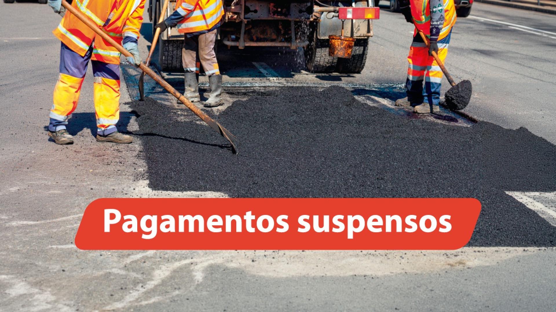 Banner horizontal com foto em ambiente externo mostrando trabalhadores com uniformes de alta visibilidade realizando serviço de pavimentação asfáltica em via pública, utilizando ferramentas manuais para espalhar o asfalto enquanto um caminhão opera ao fundo; na parte inferior da imagem, há uma faixa vermelha com o texto em destaque "Pagamentos suspensos". 