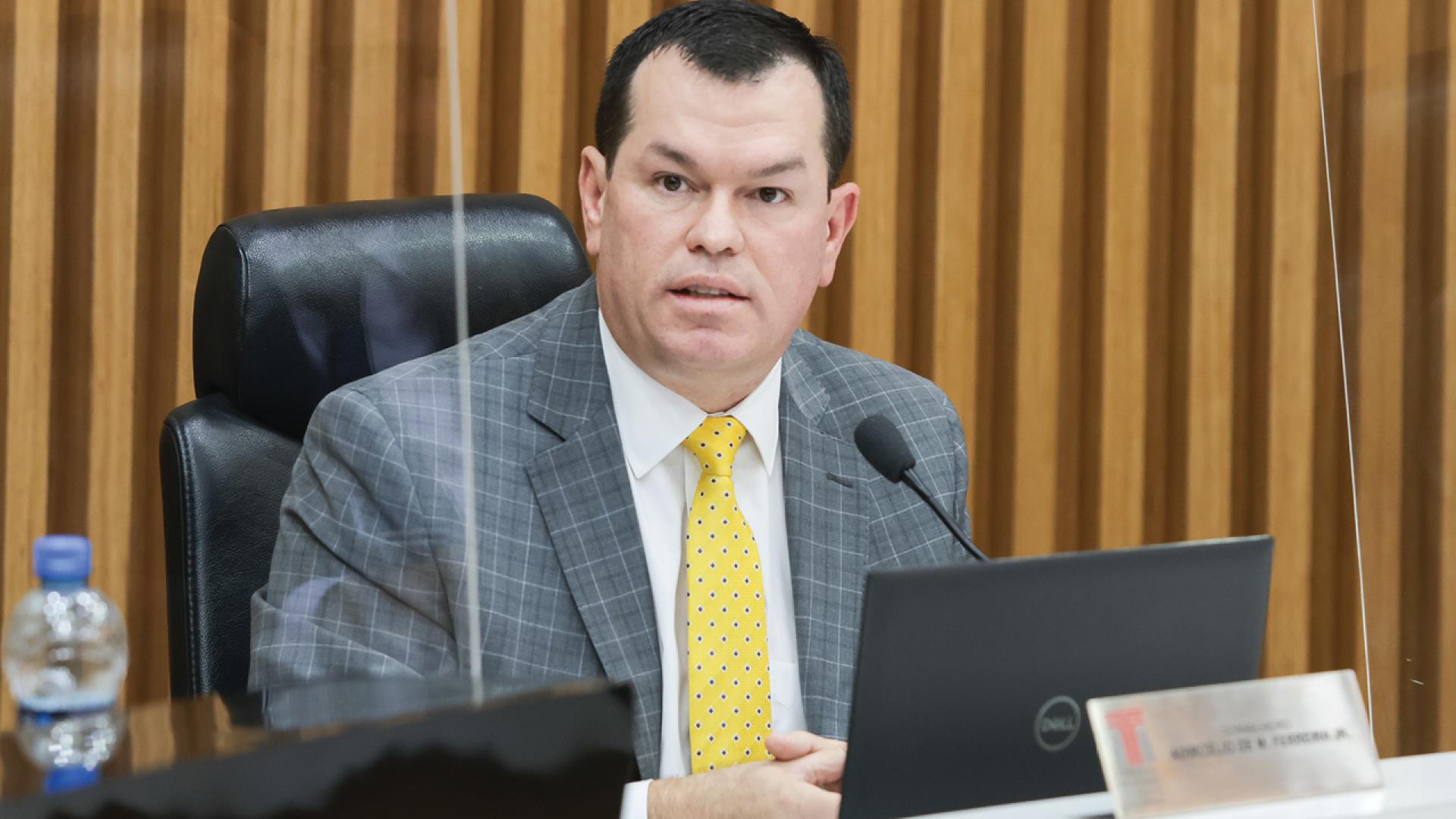 Foto horizontal do conselheiro Adircélio de Moraes Ferreira Júnior durante sessão plenária. Ele é um homem branco, de cabelos curtos e escuros. Veste terno e gravata e está sentado diante da bancada circular no Plenário. Ao fundo, painel amadeirado. À sua frente, notebook e microfone.