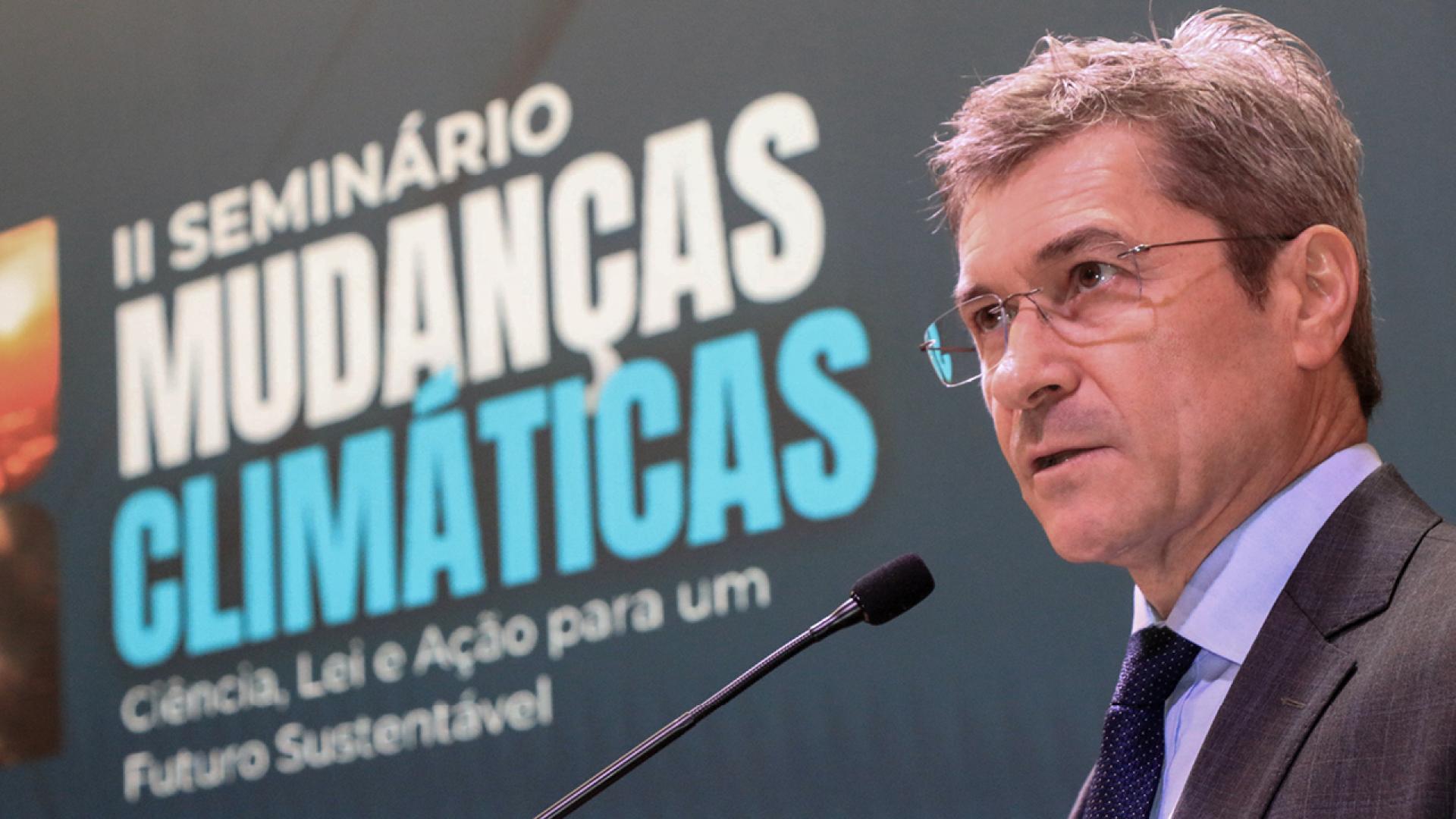 Foto do conselheiro José Nei Ascari discursando no púlpito. Ele é um homem branco, de cabelos grisalhos. Veste terno e gravata e usa óculos de grau. Ao fundo, banner com o título do evento, “segundo Seminário Mudanças Climáticas: ciência, lei e ação para um futuro sustentável”.