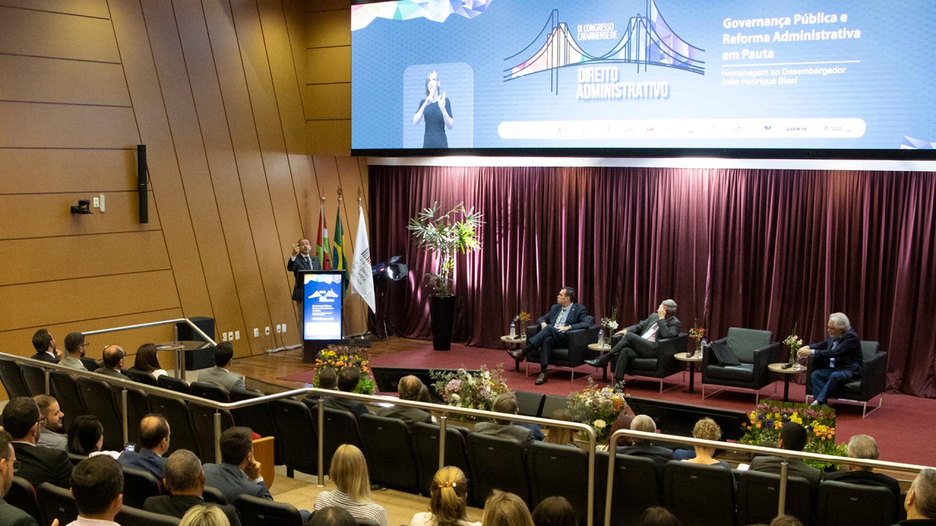 Banner horizontal com foto que mostra parte da plateia, de costas, assistindo a um dos painéis do congresso. Há várias pessoas sentadas em poltronas do auditório, lado a lado. No palco, entre os presentes, os desembargadores Hélio Pereira e João Henrique Blasi, o conselheiro Adircélio Ferreira Júnior e o presidente do Idasc, José Cristóvam. Ao fundo, no telão, é exibida a identidade visual do evento.  