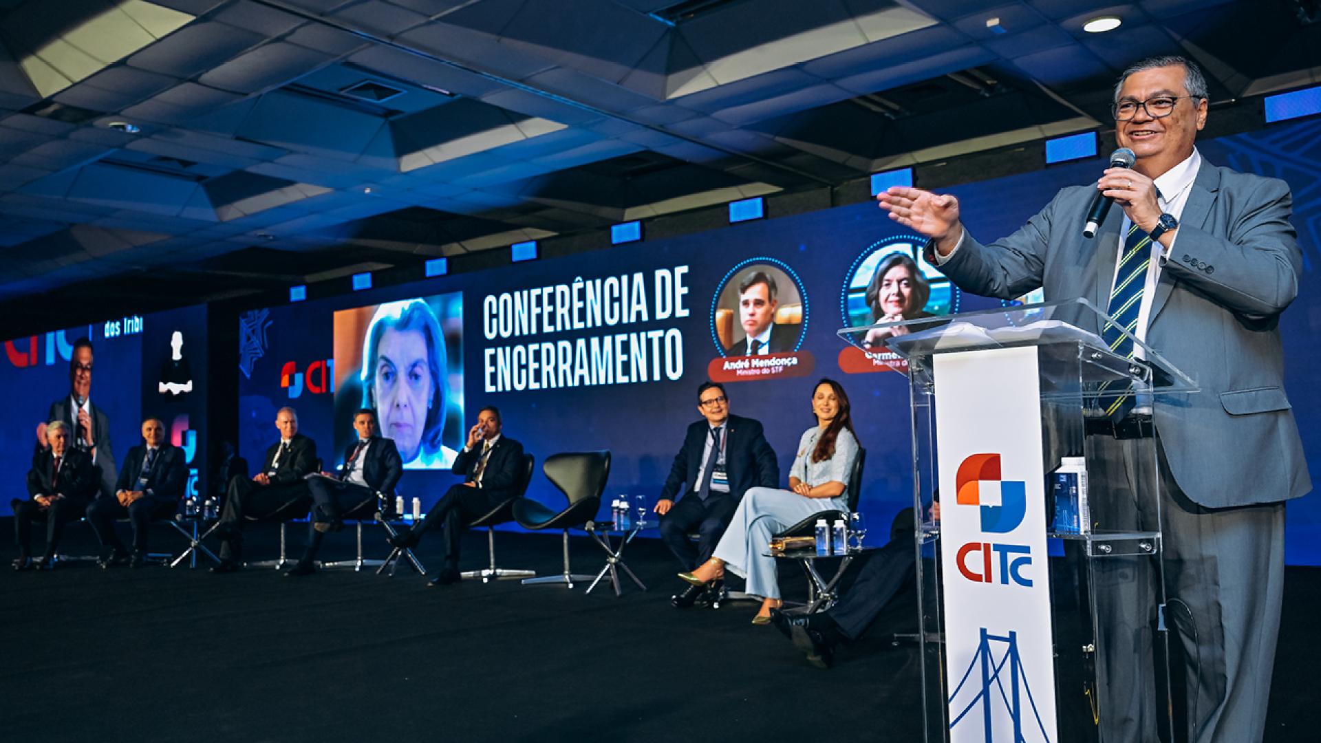 Foto do palco do IV CITC. À direita, no púlpito, o ministro do STF Flavio Dino discursa. Ao funo, telão com imgame da ministra Carmen Lucioa e os dizeres Conferencia de Encerramento. No palco ainda estão os presidentes do IRB, Atricon , TCE/SC r o ministro André Mendonça.