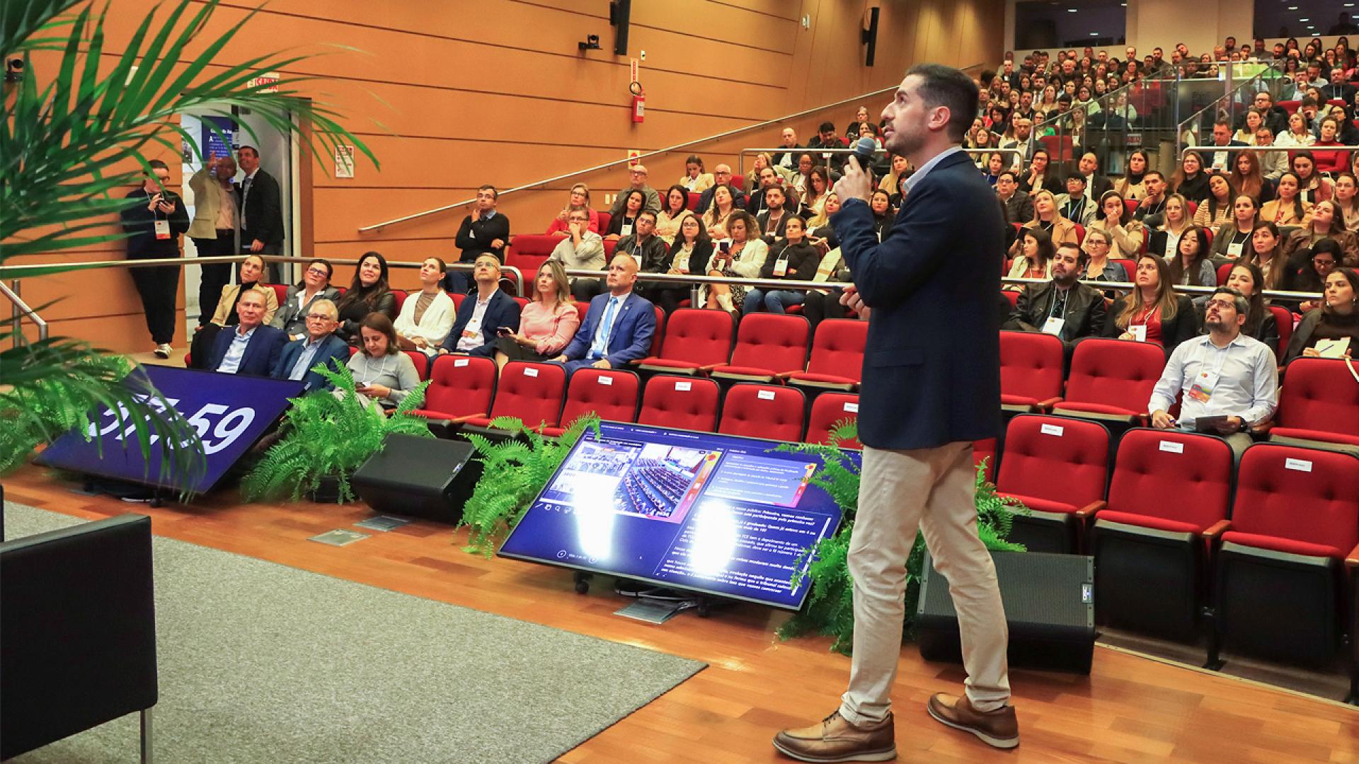 Foto mostra um homem em primeiro plano à direita, sobre um palco, olhando para o lado esquerdo. Ao fundo, há um auditório com pessoas olhando para o palco onde está esse homem