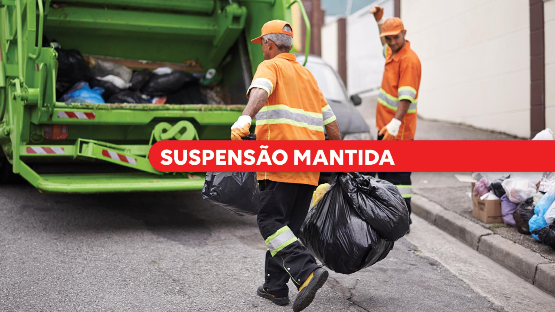 Banner horizontal com foto de trabalhadores da coleta de resíduos urbanos. Em primeiro plano, um funcionário uniformizado com roupa laranja e faixas refletivas carrega sacos de lixo em direção a um caminhão de coleta verde. Ao fundo, outro trabalhador sinaliza próximo ao veículo, enquanto sacos de resíduos aparecem dispostos na calçada. Sobre a imagem, uma faixa vermelha em destaque exibe o texto "Suspensão mantida". 