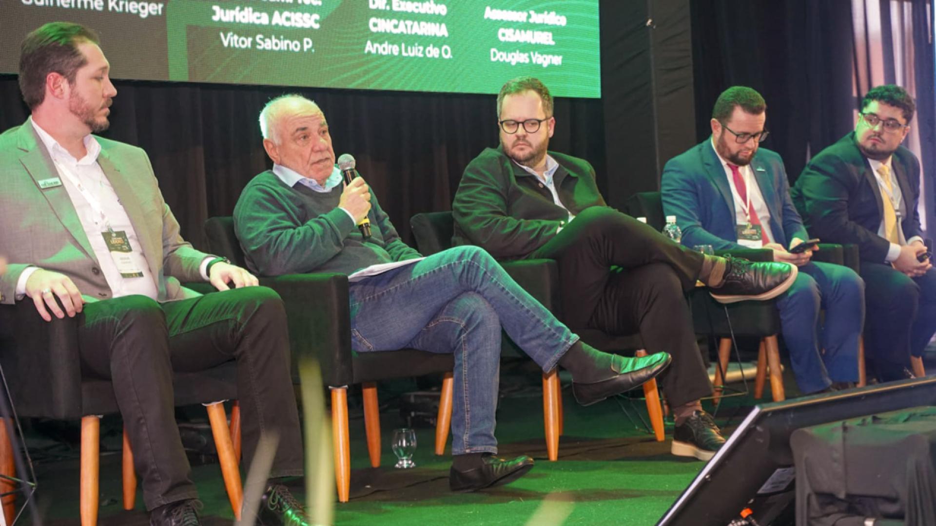 Imagem mostra cinco homens sentados em cadeiras em um palco. O segundo homem da esquerda para a direita, o conselheiro Luiz Eduardo Cherem, está falando ao microfone