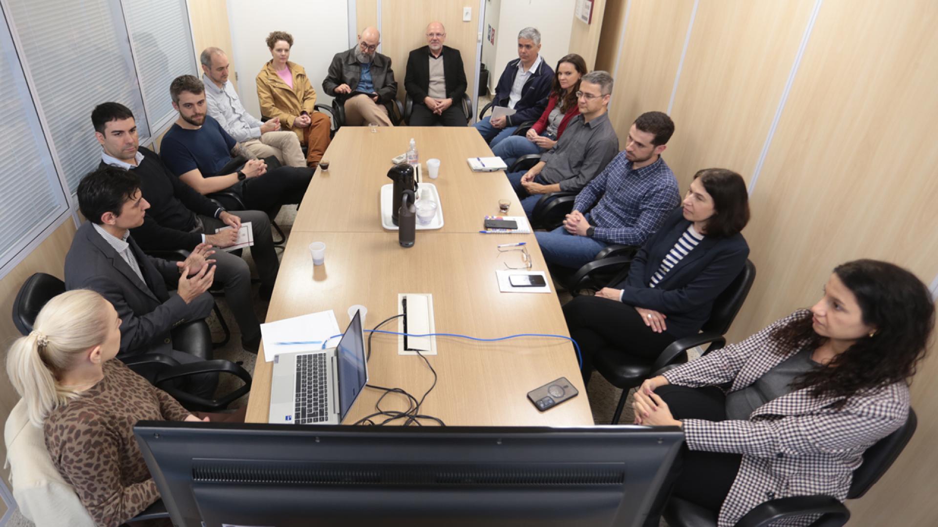Foto de várias pessoas sentadas diante de uma mesa de reuniões durante apresentação dos novos módulos do SEI. Entre os presentes, o diretor de Tecnologia da Informação, Rafael Queiroz, que fala a outros integrantes do TCE/SC. Sobre a mesa, há notebook, papéis e celulares.