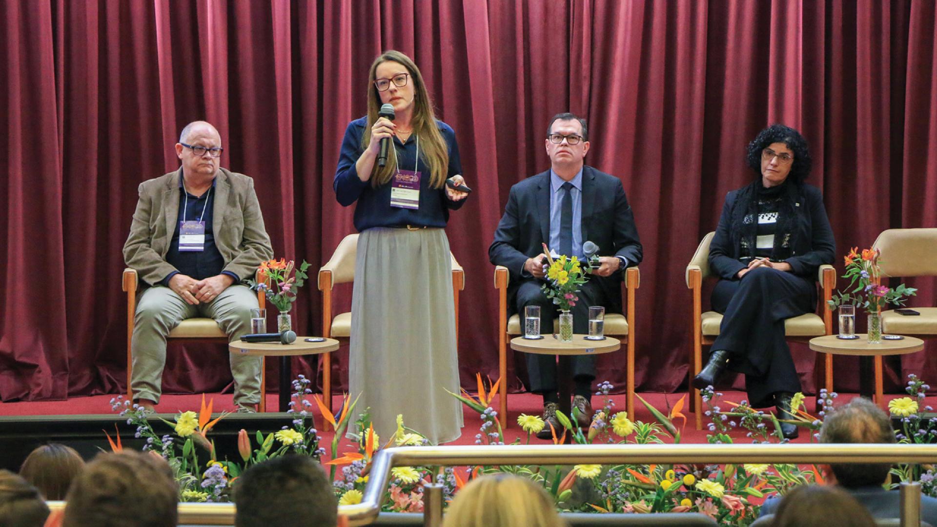 Banner horizontal com foto da palestrante Vivian Savedra falando ao microfone durante o evento. Estão, junto com ela no palco, outras três pessoas, incluindo o conselheiro Adircélio Ferreira Júnior. O cenário é composto por cortinas bordôs e plantas decorativas.