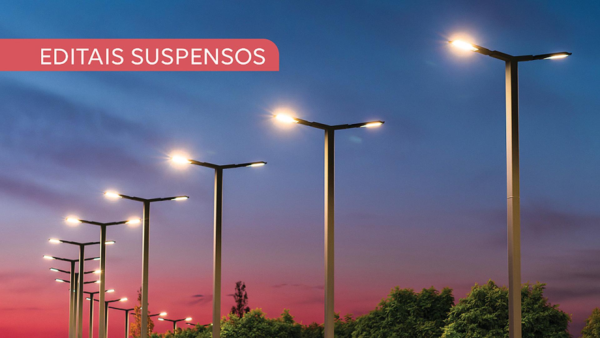 Imagem mostra vários postes de iluminação em perspectiva. O céu ao fundo vai em degradê do azul para o roxo. No alto, à esquerda, há a inscrição Editais Suspensos em branco sobre uma tarja vermelha