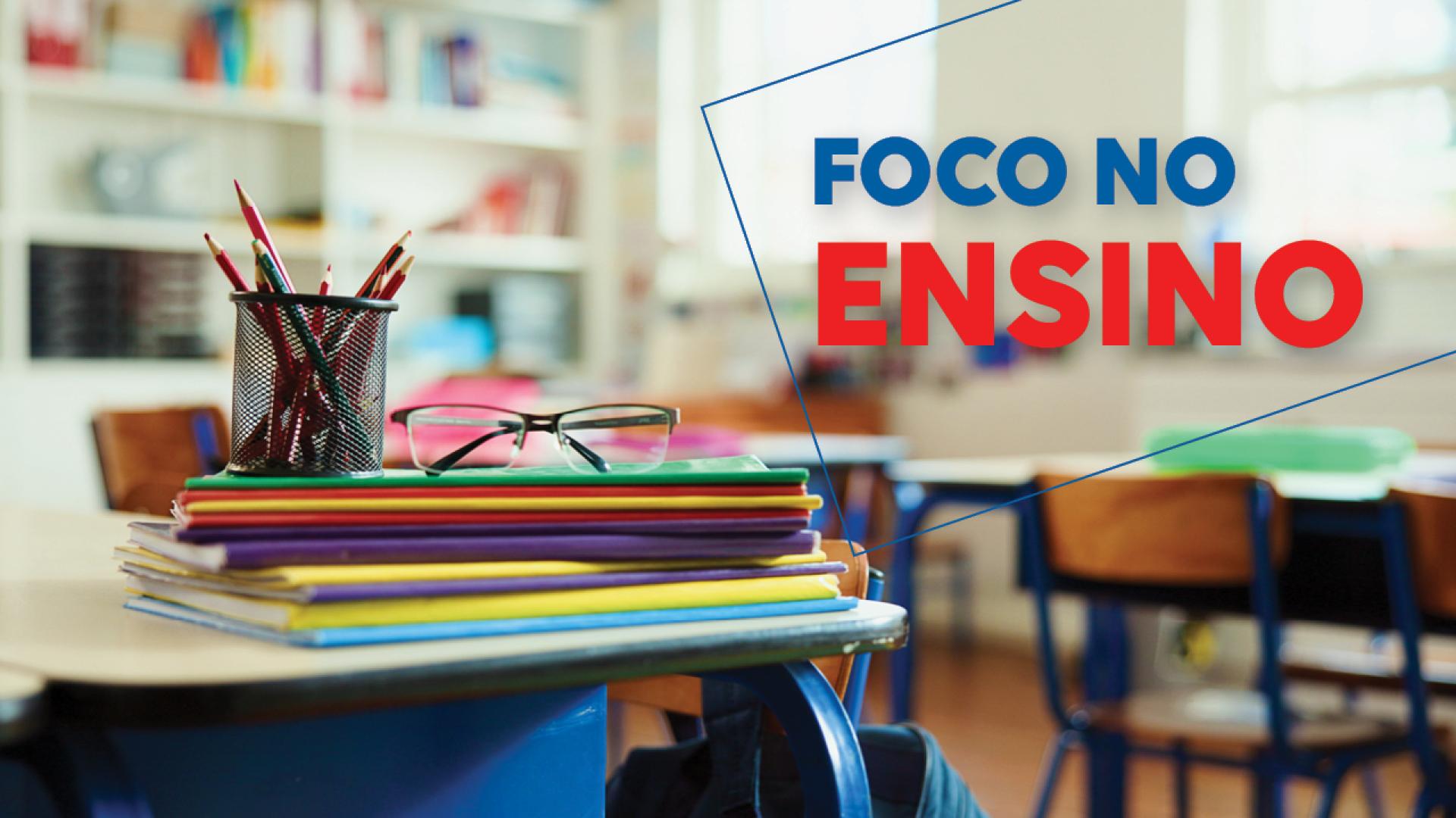 Banner horizontal com foto de materiais escolares empilhados sobre cadernos em uma mesa. Há um porta-lápis e óculos de grau, cadernos e livros. Sobre a foto, o texto "Foco no ensino", em fonte colorida.