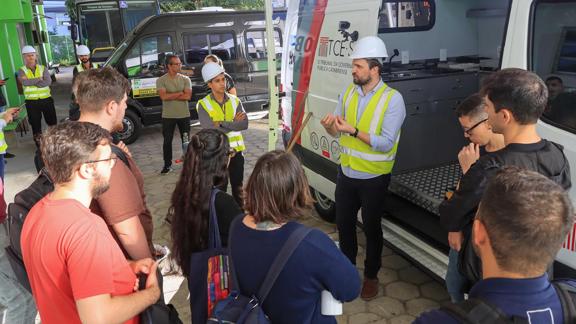 Foto horizontal, no pátio do IFSC, em que aparecem os técnicos do TCE/SC, trajados com colete amarelo e capacete branco, diante do caminhão laboratório de obras rodoviárias, passando instruções às pessoas que assistem, de pé. 