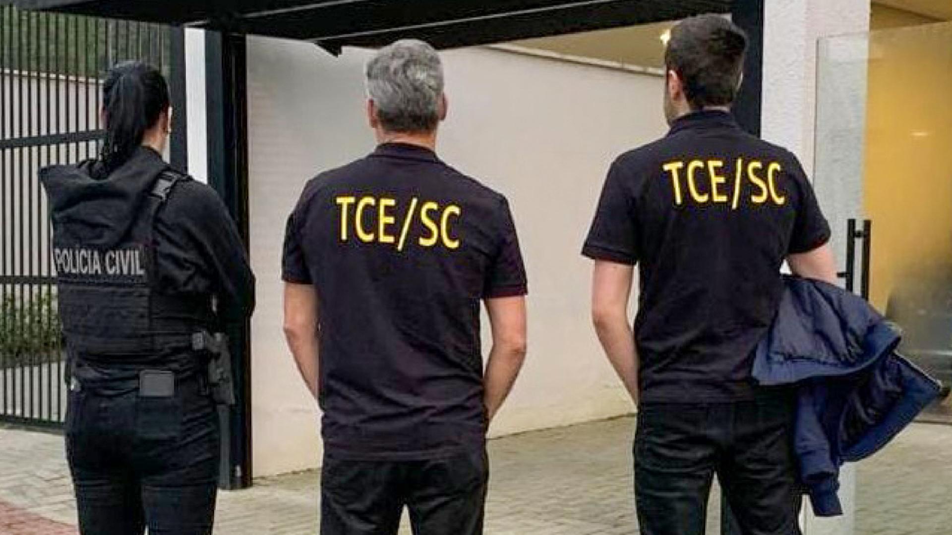 Fotografia mostra três pessoas vestidas de roupas escuras e de costas. À esquerda há uma mulher que usa um colete com as inscrições da Polícia Civil. Os outros dois são homens e estão com uma camisa escrita TCE/SC. 