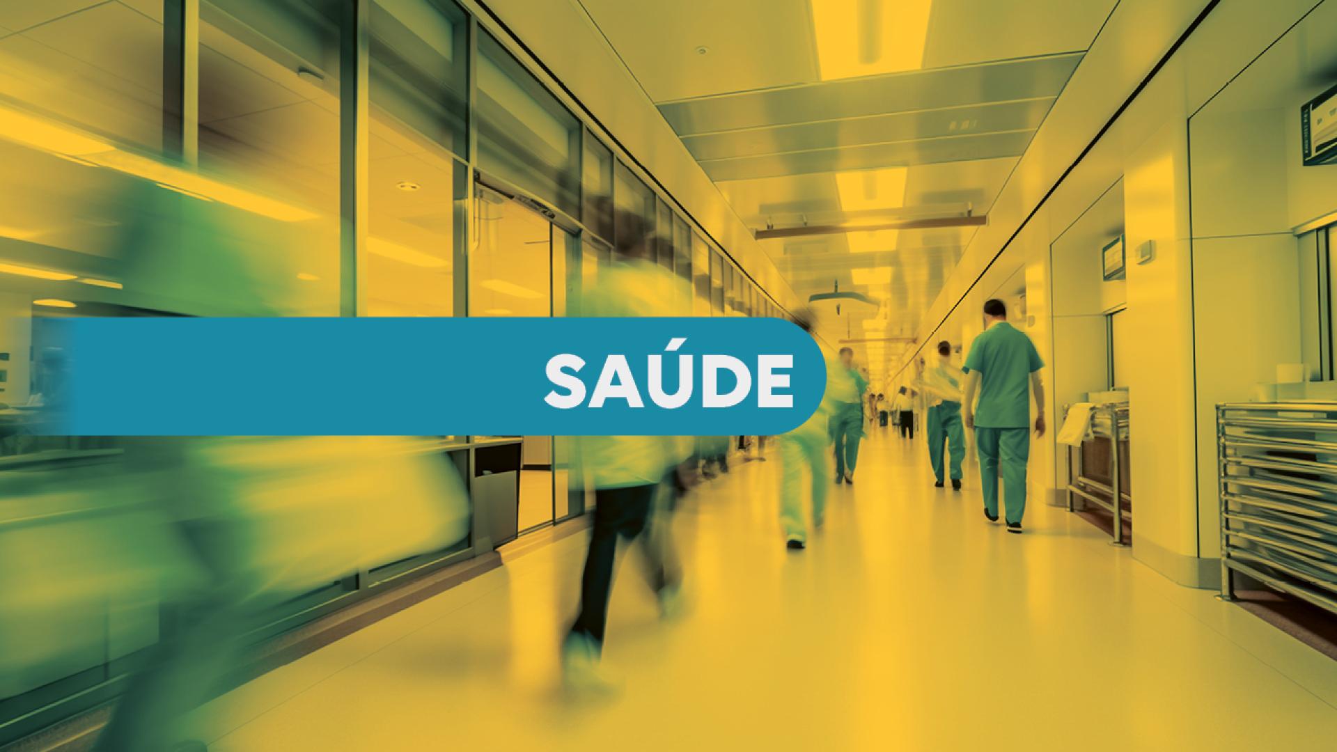 Banner horizontal. Imagem desfocada de um corredor de hospital, onde algumas pessoas caminham. A imagem tem tons amarelos e as roupas das pessoas em azul. Na parte central uma faixa azul sobre a qual, em letras brancas, está escrita a palavra SAÙDE.