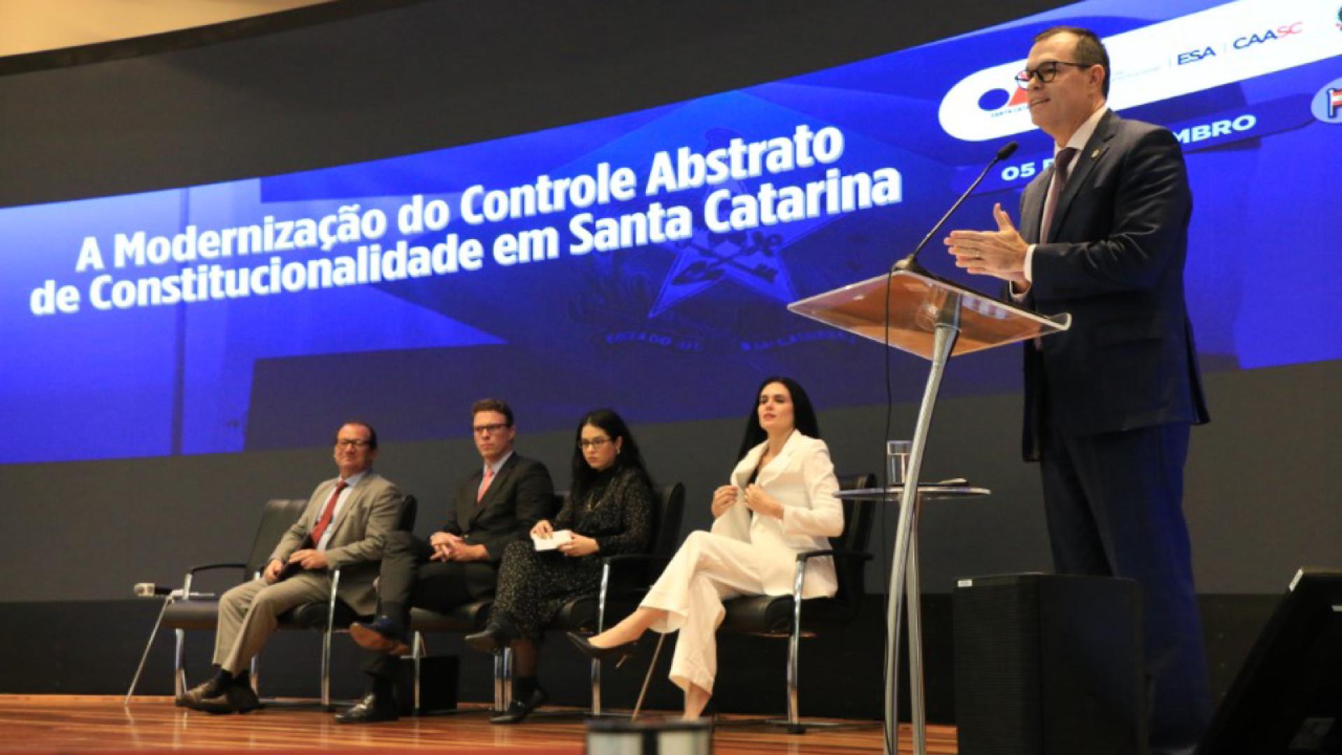 A imagem traz cinco pessoas em um palco, durante uma palestra, sendo que quatro estão sentadas a partir da esquerda para direita: dois homes e duas mulheres. De pé, no canto direito da foto, um homem, conselheiro do Tribunal de Contas de Santa Catarina Adircélio de Moraes Ferreira Júnior, faz uma apresentação em frente a um púlpito. Ele veste terno azul-escuro e está com as duas mães estendidas a frente.
