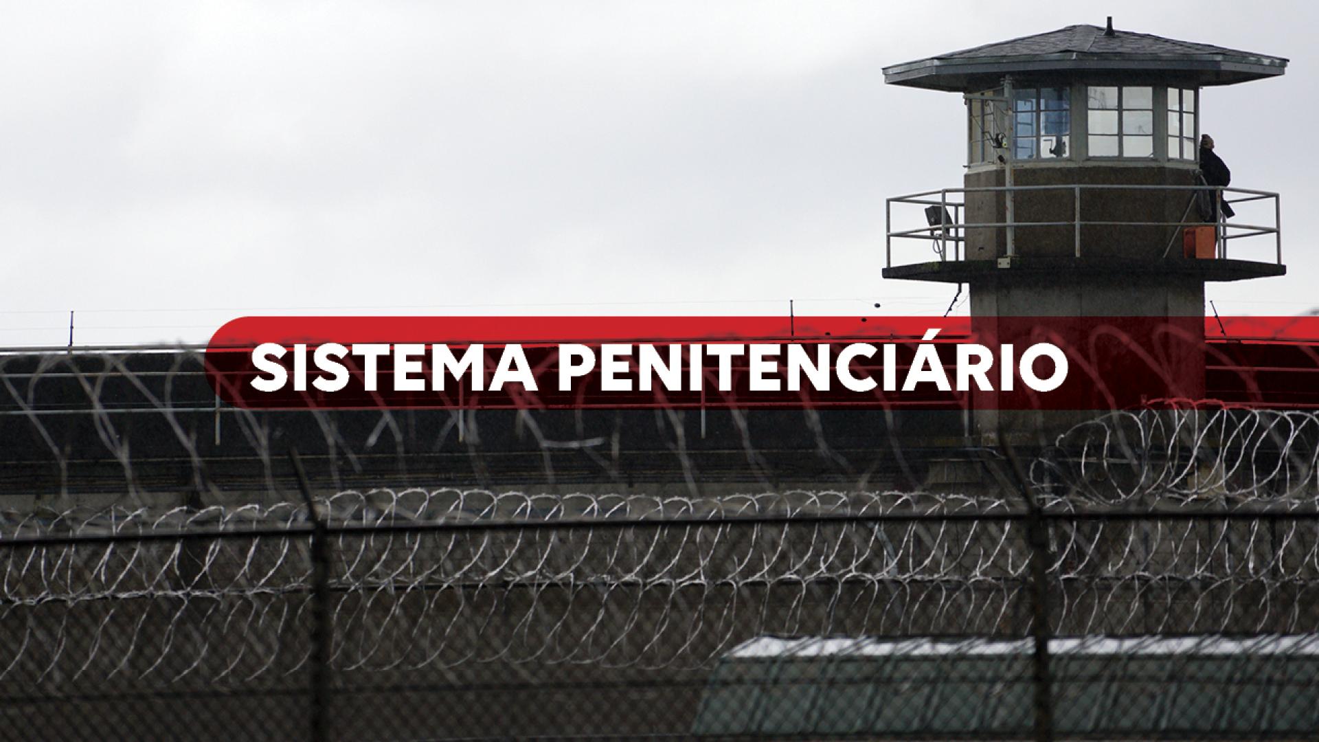 Imagem mostra o muro de uma penitenciária com uma guarita no alto, à direita. Ao centro há a expressão Sistema Prisional escrito em branco sobre um tarja vermelha.