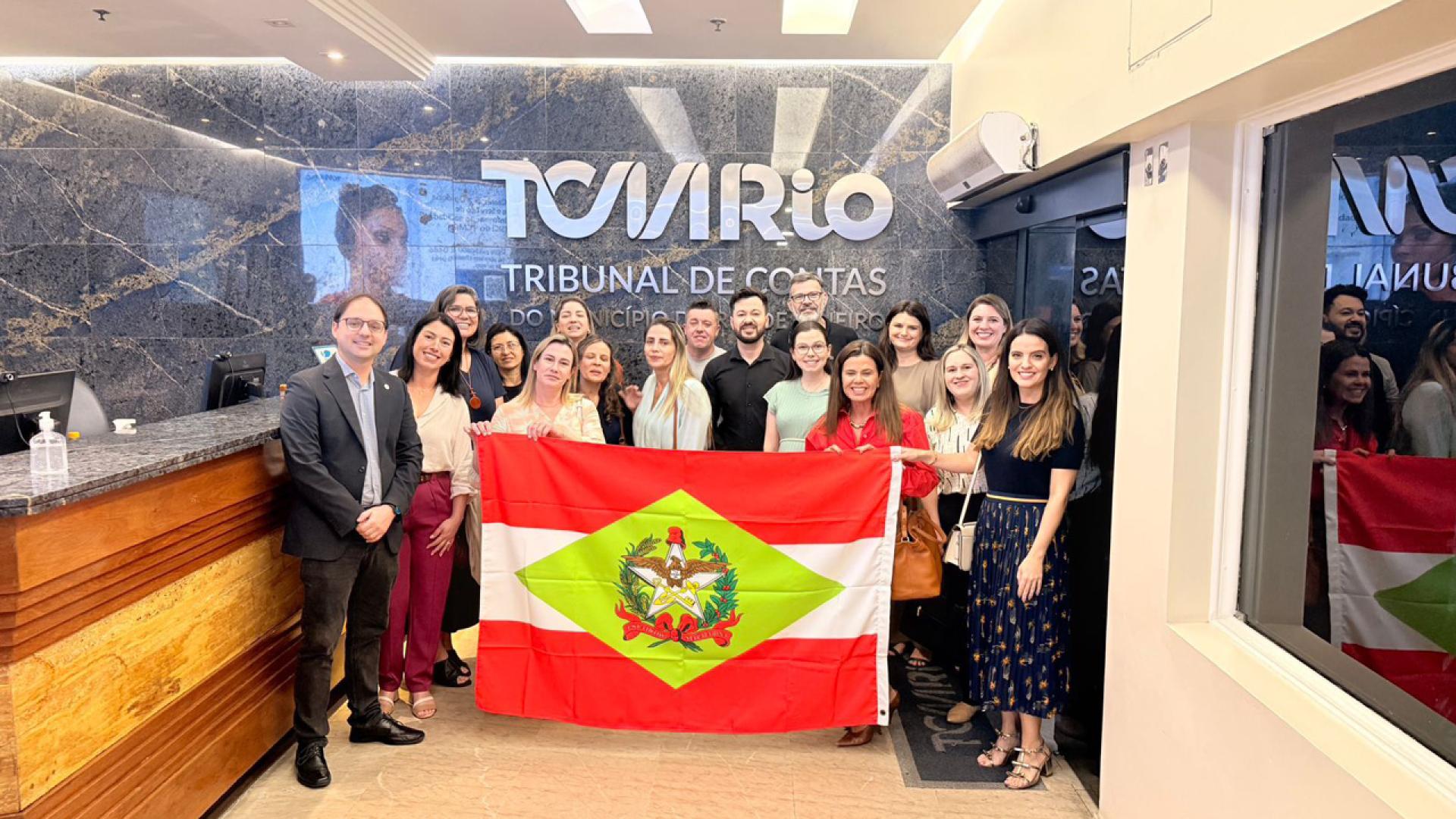 Banner horizontal com foto da comitiva catarinense segurando uma bandeira de Santa Catarina em ambiente interno no TCMRio. Entre os presentes, diretoras das escolas vencedoras e servidores do TCE/SC