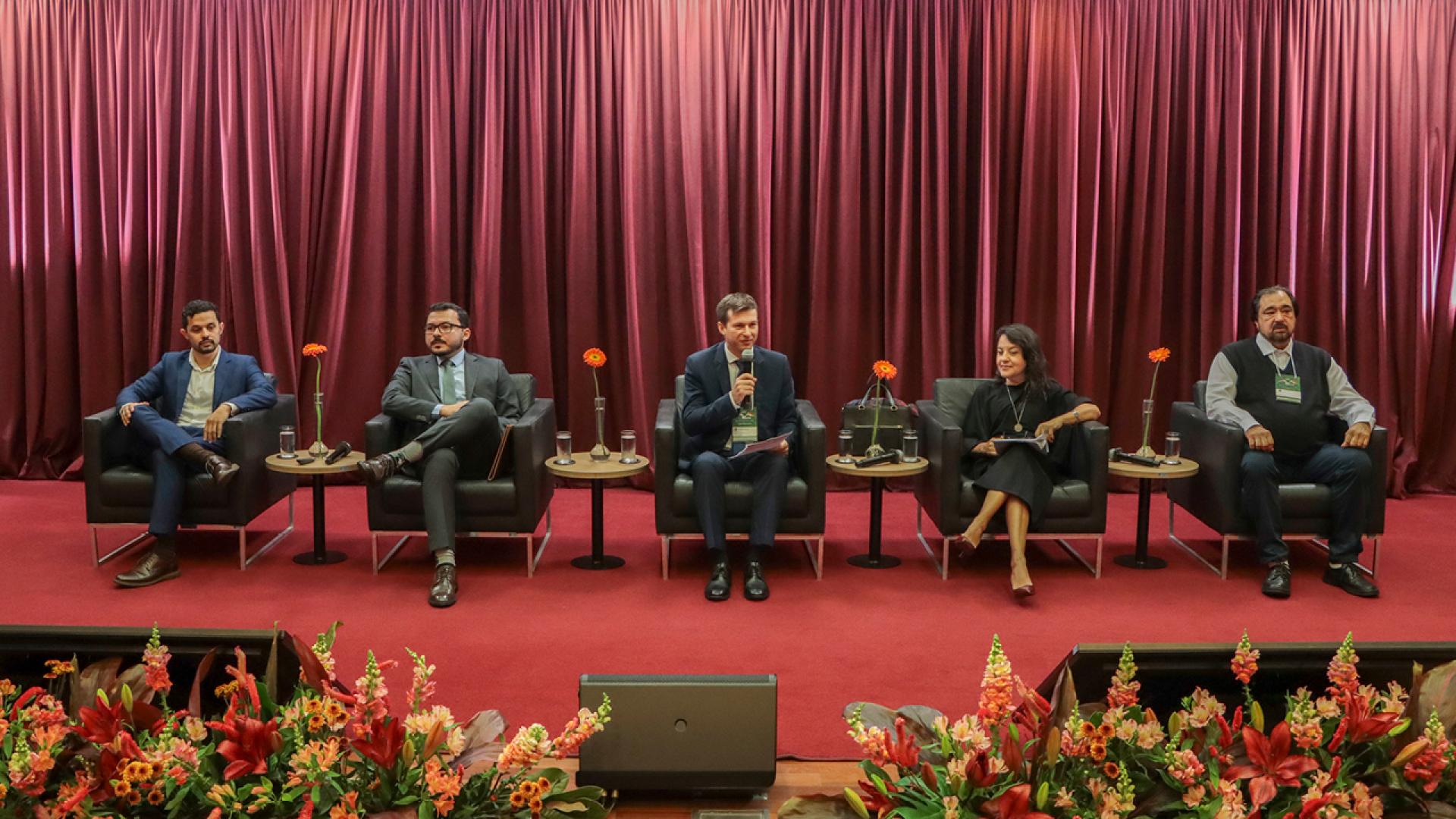 Imagem mostra cinco pessoas sentadas em cadeiras num palco. São quatro homens e uma mulher.
