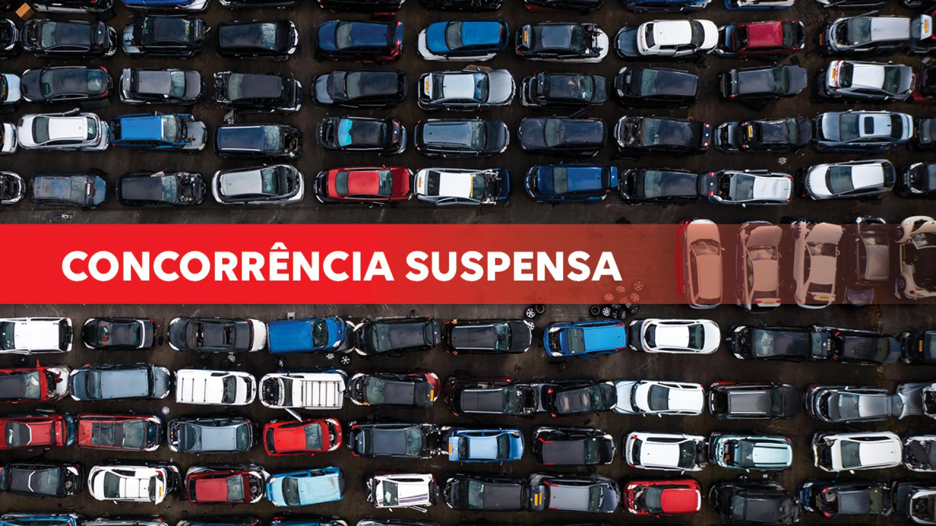 Banner horizontal com foto de grande quantidade de carros estacionados lado a lado, vistos de cima, formando várias fileiras apertadas. Os veículos têm diferentes cores, como preto, branco, vermelho, azul e cinza, e ocupam praticamente todo o espaço da imagem. Sobre a foto, uma tarja vermelha com o texto "Concorrência suspensa".