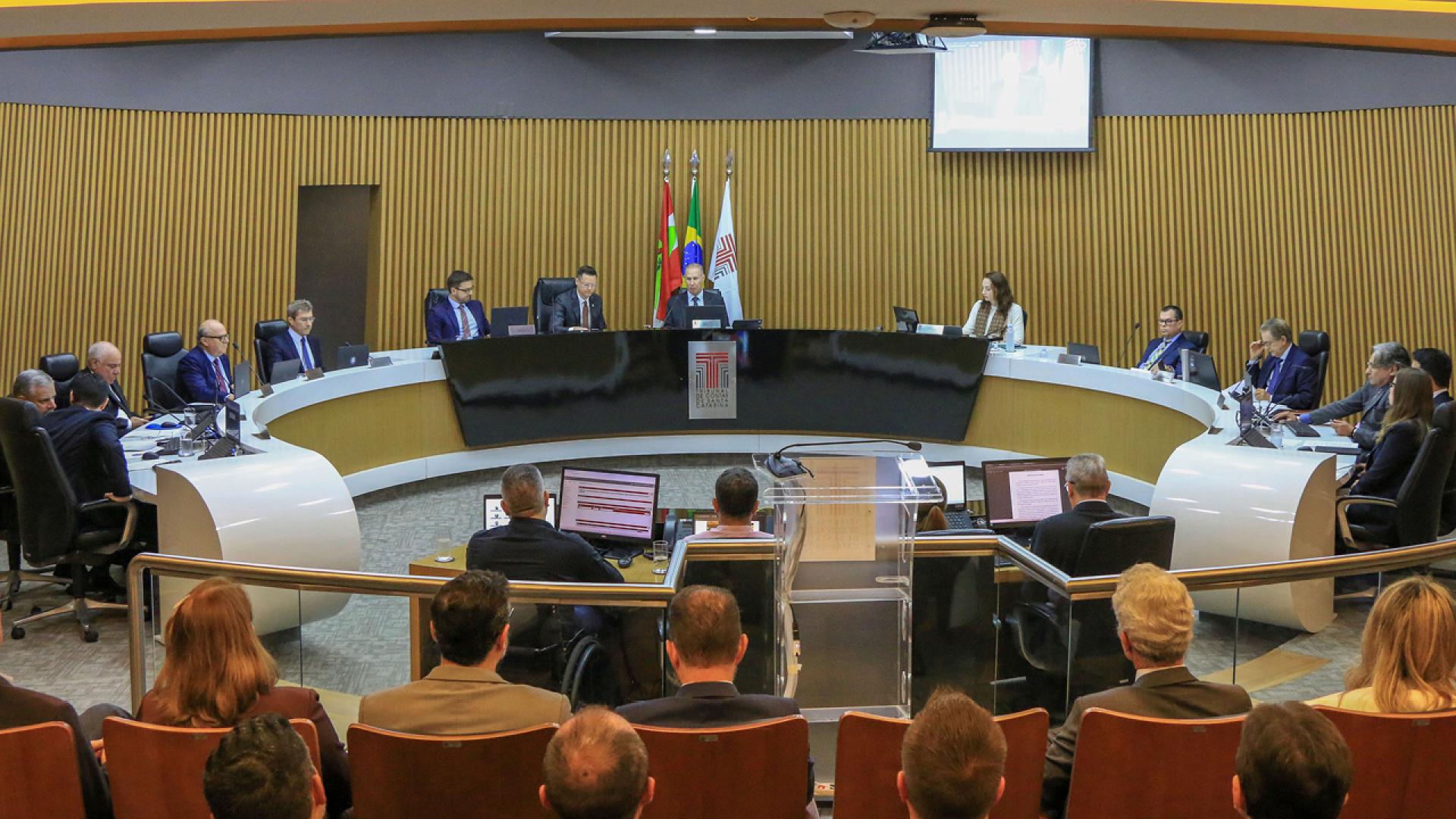 Banner horizontal com foto do plenário do TCE/SC durante sessão de apreciação das contas. Ao centro, conselheiros e conselheiros substitutos estão posicionados em uma grande mesa semicircular. No centro da composição, está a mesa diretiva da sessão, conduzida pelo presidente Herneus De Nadal e pelo relator do processo, conselheiro Luiz Roberto Herbst. Também participam da sessão procuradores MPTC/SC e representantes do governo estadual, entre eles o secretário da Fazenda, Cleverson Siewert. 