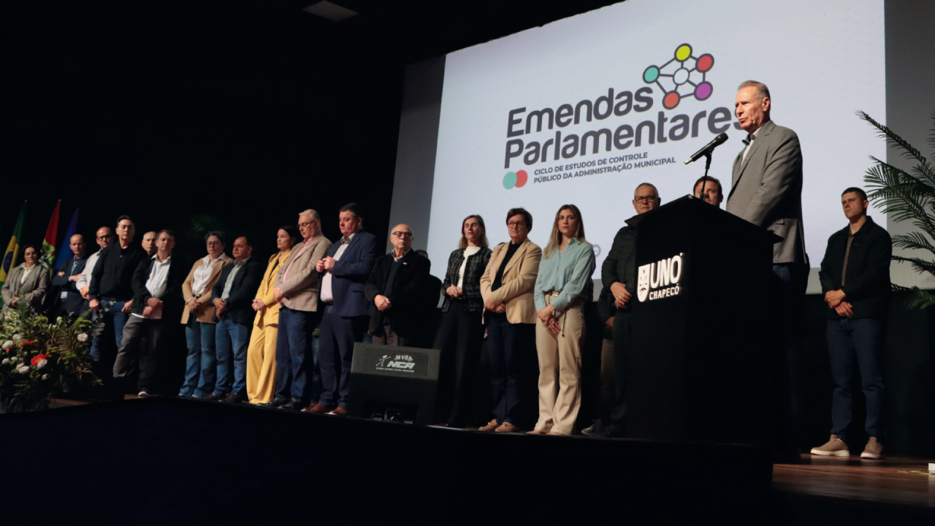 Banner horizontal com foto da comitiva do TCE/SC posicionada no palco do auditório. No púlpito, o presidente Herneus discursa. Ao fundo, painel com logo do evento Emendas Parlamentares. A iluminação do auditório é em tons escuros.