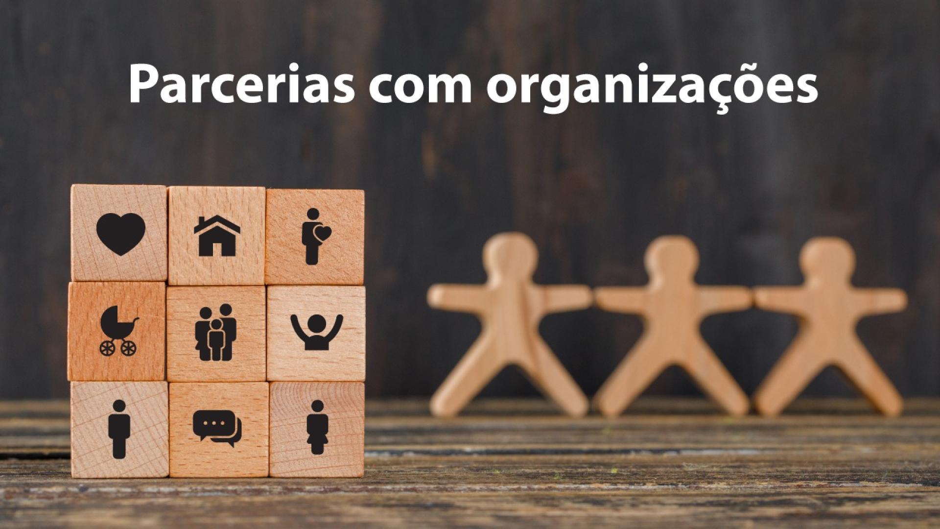 Banner horizontal com imagem ilustrativa de um cubo de madeira com ícones que representam áreas ou temas sociais, como coração, casa, acessibilidade, família, inclusão, diálogo e pessoas. À direita, aparecem três figuras de madeira estilizadas, de mãos dadas. Sobre a foto, na parte superior, lê-se "Parcerias com organizações". 