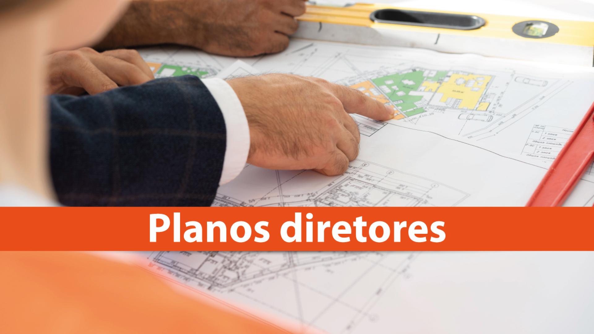 Banner horizontal com foto de pessoas analisando plantas arquitetônicas sobre uma mesa. Em destaque, uma mão aponta para uma área colorida do projeto. Na parte inferior, em tarja alaranjada, o texto “Planos diretores”, em fonte branca. 