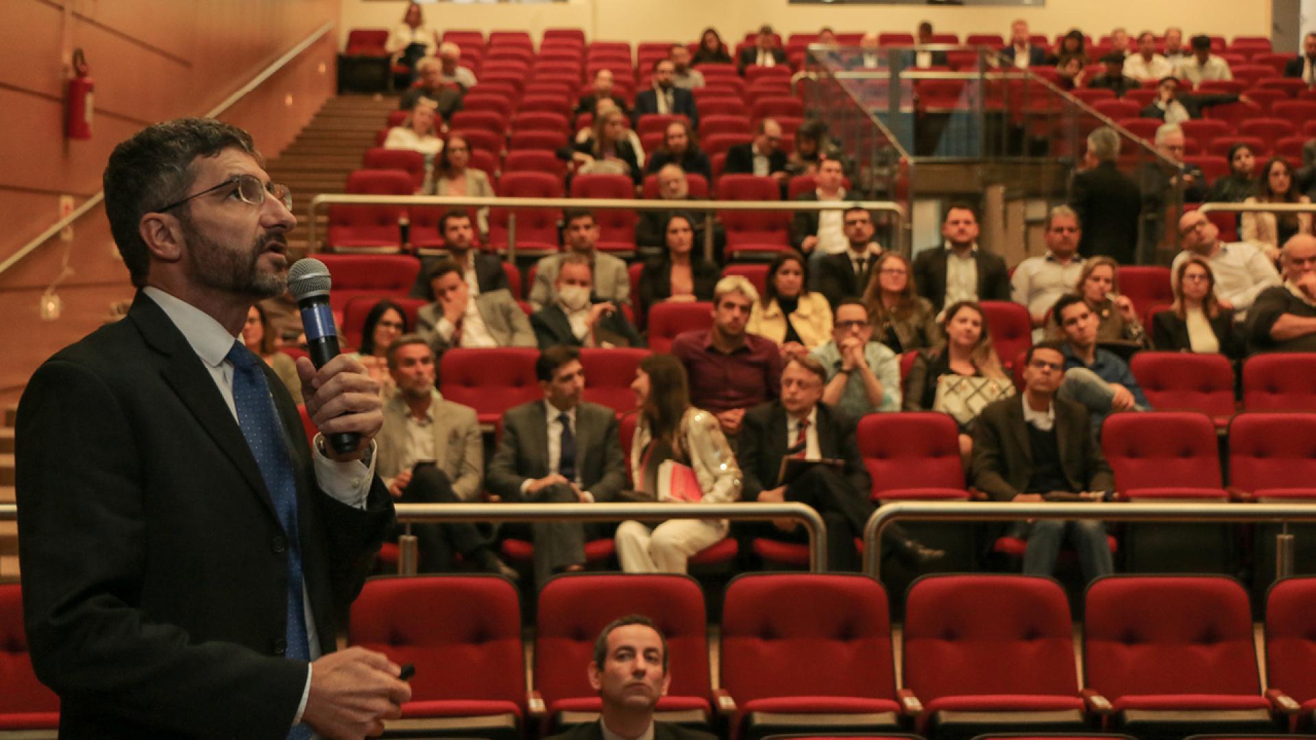 Foto do auditório do TCE, a partir do palco em direção à plateia, com o público ao fundo. Em primeiro plano, à esquerda, o palestrante Azor El Achkar olhando para a direita, segurando o microfone. Azor é branco, de barba, e está de terno preto e gravata azul.