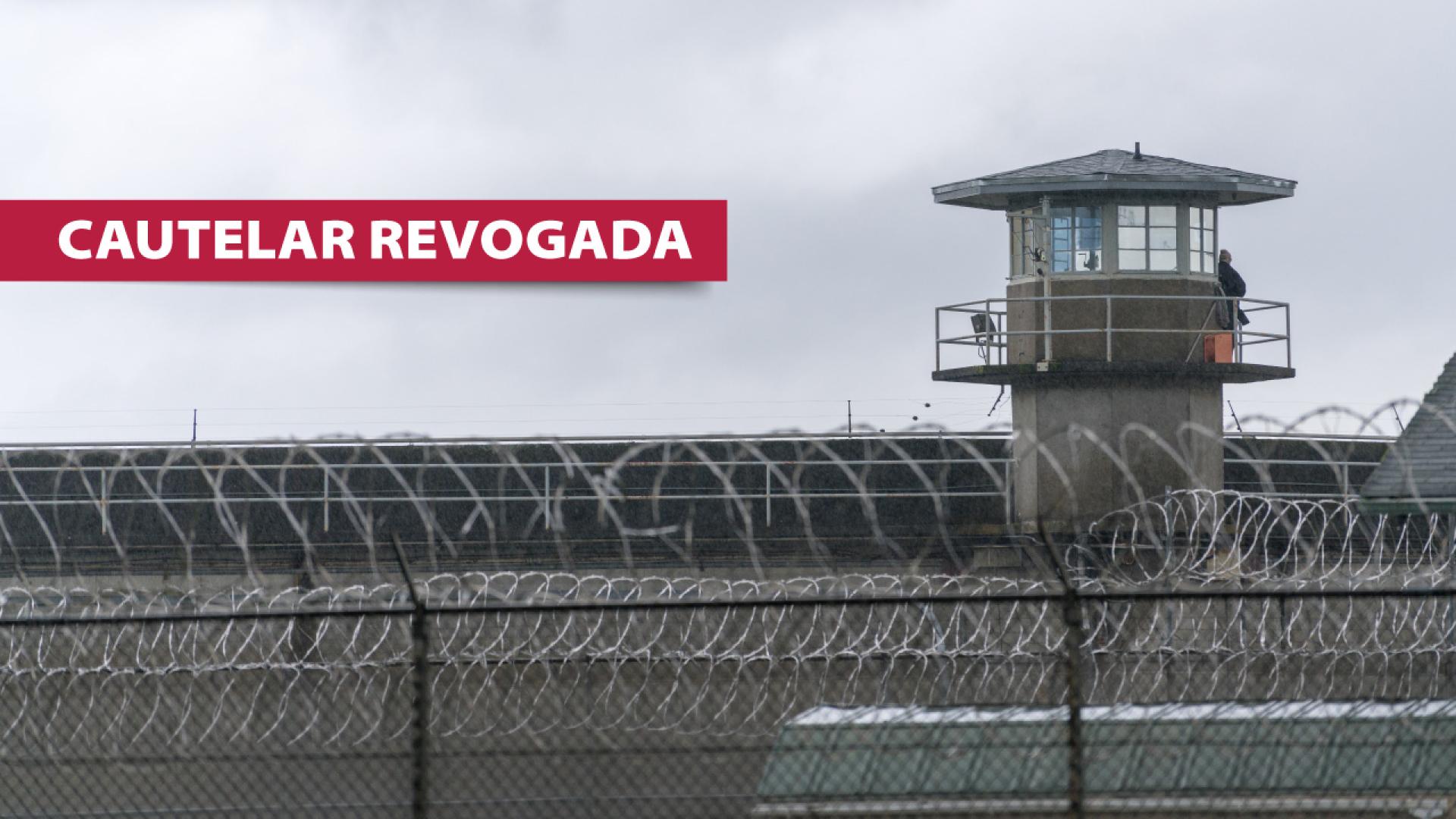 Imagem mostra o muro de uma penitenciária e uma torre de vigilância. À esquerda, escrito em branco sobre tarja vermelha, está escrito “cautelar revogada”