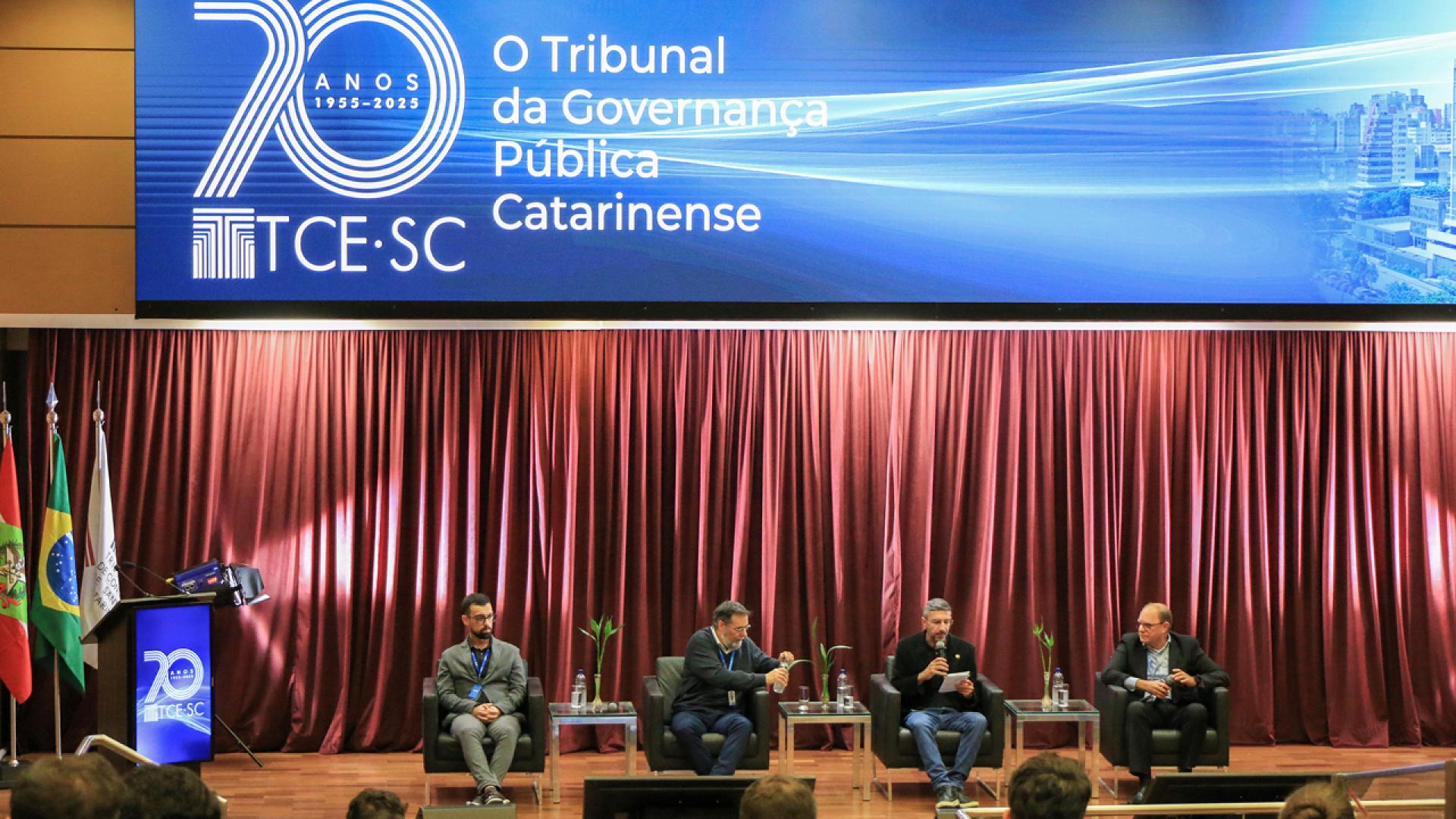 Foto horizontal do palco do TCE/SC. Os 4 integrantes da roda de conversa estão sentados lado a lado. No fundo, na parte superior o telão mostra a logo do TCE/SC.