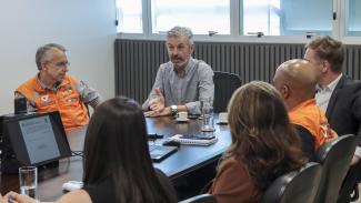 Imagem mostra seis pessoas sentadas ao redor de uma mesa. Quatro delas estão de costas. Aparecem de frente dois homens. À esquerda, um homem com cabelos grisalhos vestindo um colete alaranjado. Mais ao centro, um homem de cabelos grisalhos e barba vestindo uma camisa cinza