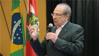 Foto do conselheiro aposentado Sebastião Helvécio, proferindo palestra no auditório do TCE/SC. Ele é um homem branco, de cabelos grisalhos. Veste terno e gravata, usa óculos de grau e está em pé, falando ao microfone. Atrás dele, aparecem as bandeiras do Brasil e de Santa Catarina. 