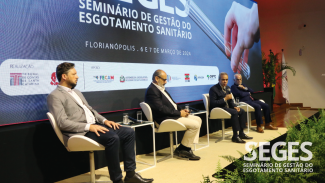 Foto dos quatro painelistas durante o evento. Da esquerda para a direita: o presidente da Assemae/SC, Osvalcir Peters, o diretor-executivo da ABCON, Percy Neto, o presidente da Atricon, conselheiro Edilson de Sousa Silva e o presidente da Casan, Edson Moritz. Ambos estão sentados em poltronas no palco do auditório. Ao fundo, no telão, projeção do logo do evento. 