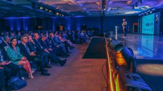 Foto horizontal de uma das palestras. À esquerda, várias pessoas com fones de ouvido, sentadas em cadeiras, lado a lado. À frente delas, à direita da foto, o palestrante, no palco, falando aos demais. O ambiente tem luzes azuis.