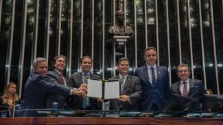 Banner horizontal com foto dos integrantes da mesa de autoridades no Senado segurando um documento. Há seis pessoas, lado a lado, que vestem terno e gravata. Entre os presentes, o presidente do Senado, Davi Alcolumbre, o presidente do TCU, ministro Vital do Rêgo, e o presidente da Atricon, Edilson Silva. 