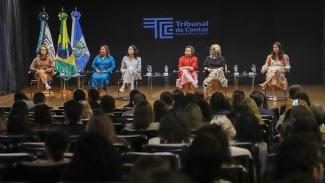 Banner horizontal com foto da plateia acompanhando um painel de debates realizado no TCE-RJ. No palco, seis mulheres estão sentadas em poltronas dispostas lado a lado, com mesas de apoio e copos de água à frente. Entre as palestrantes, está a conselheira substituta do TCE/SC Sabrina Iocken. 